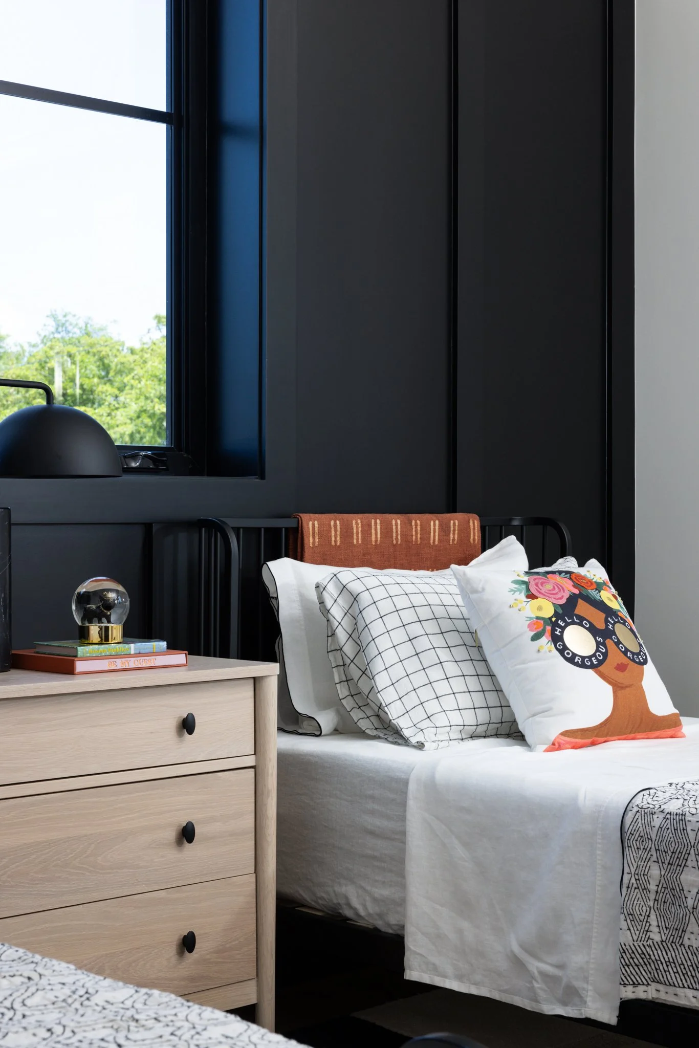 A modern guest kids bedroom interior with a bed, throw pillows, a oak wooden nightstand with books and a decorative lamp, a large window with outdoor greenery, and dark-colored wall paneling.