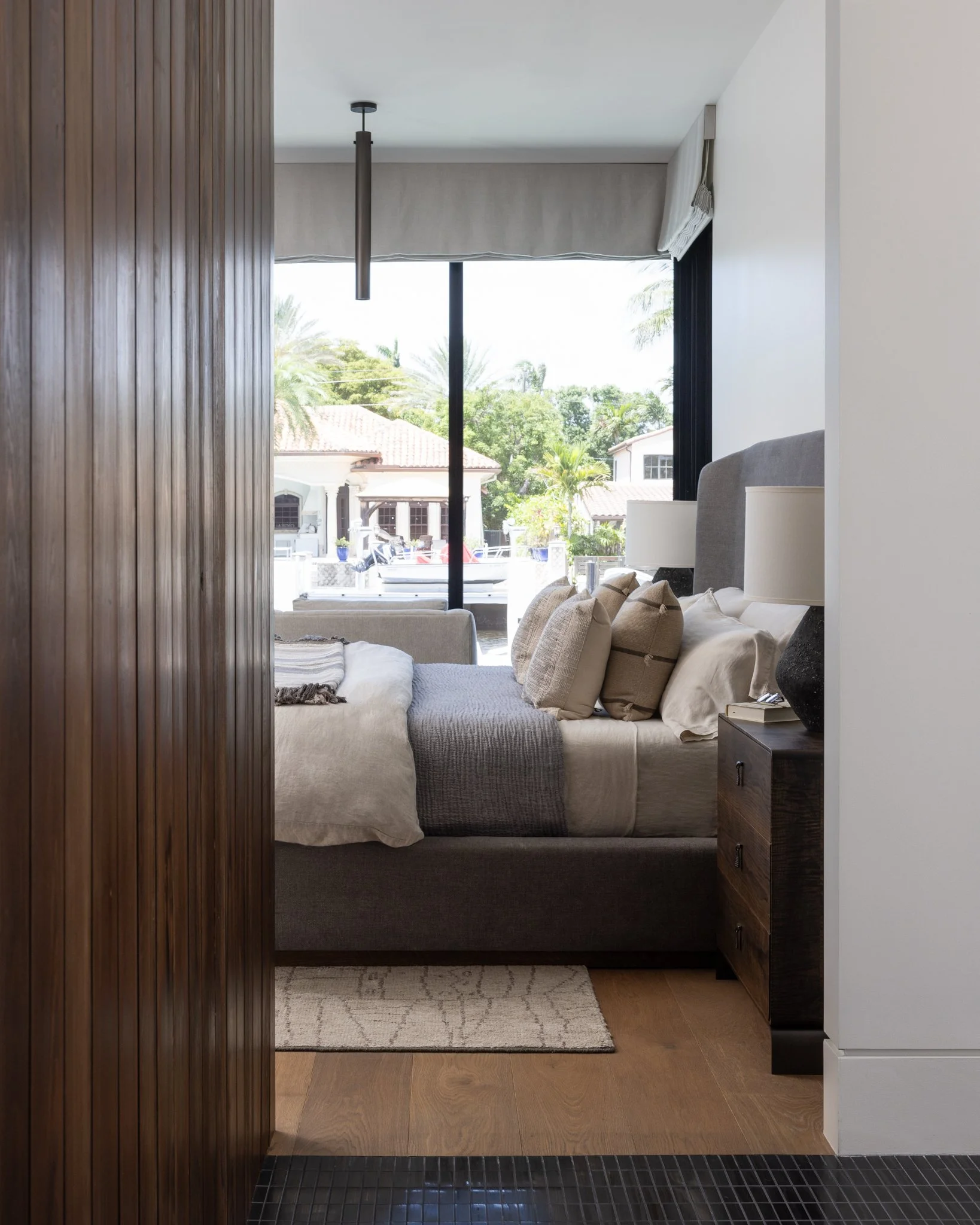 View of a cozy bedroom with a custom made bed, multiple pillows, and a gray throw blanket, facing a large window revealing a sunny waterfront outdoor scene with palm trees and houses, and custom made nightstand by joe wolf of wolf wood co. 