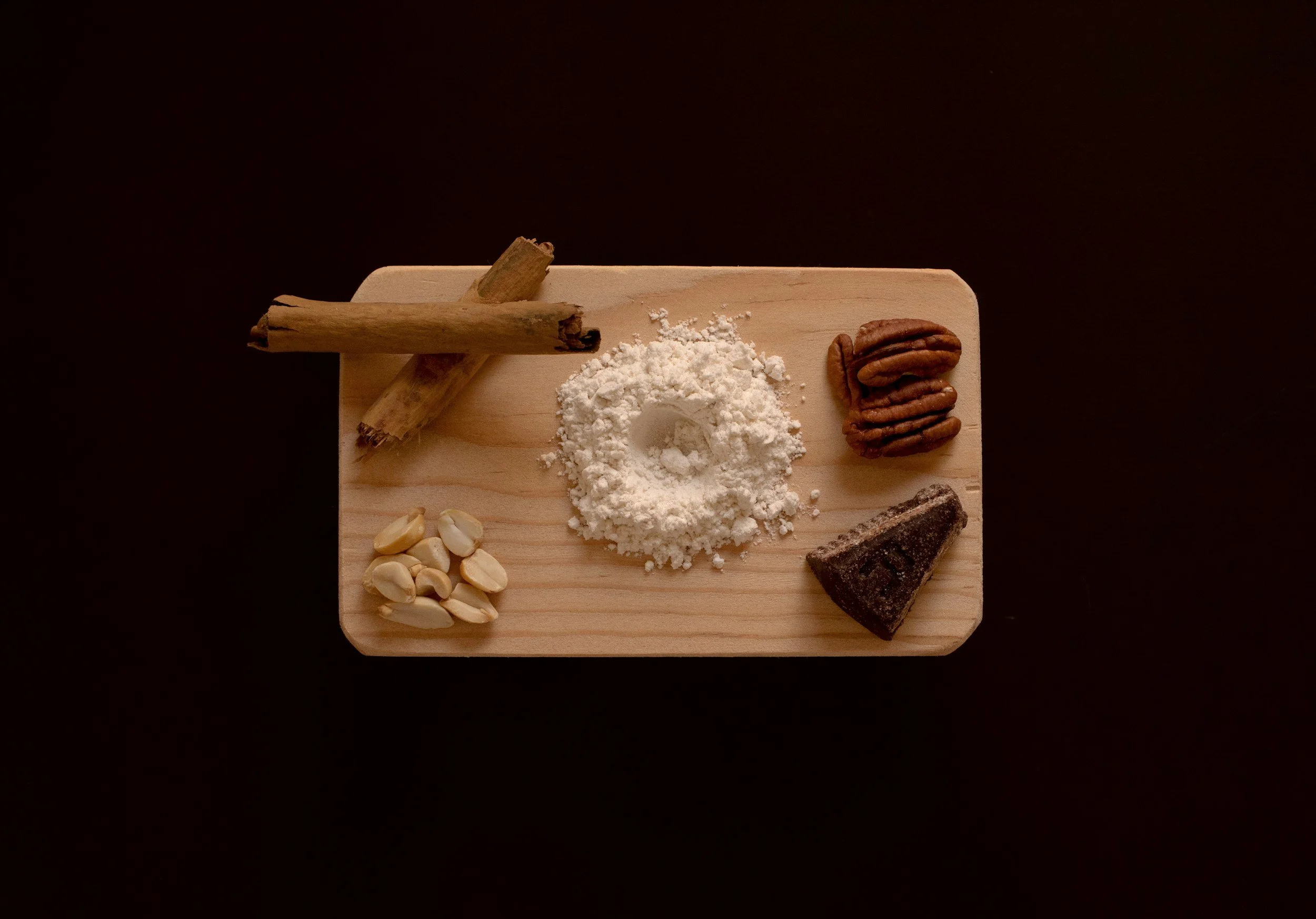 A wooden board with garlic cloves, crushed garlic, flour, pecans, a broken piece of chocolate, and cinnamon sticks on a dark background.