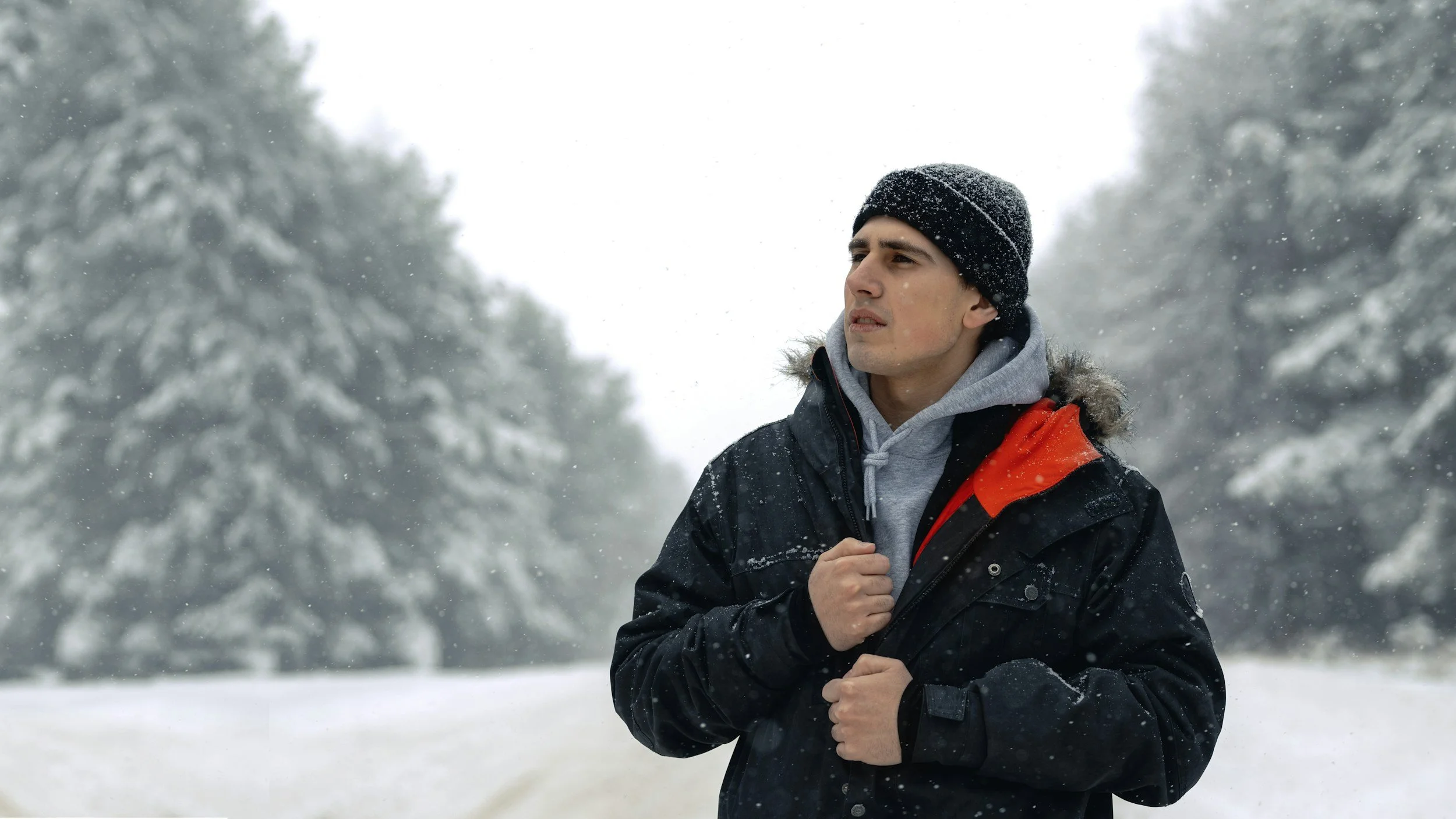 A man wearing a black beanie, gray hoodie, and black winter coat with a red lining, jogging in a snowy outdoor landscape with snow-covered trees in the background.