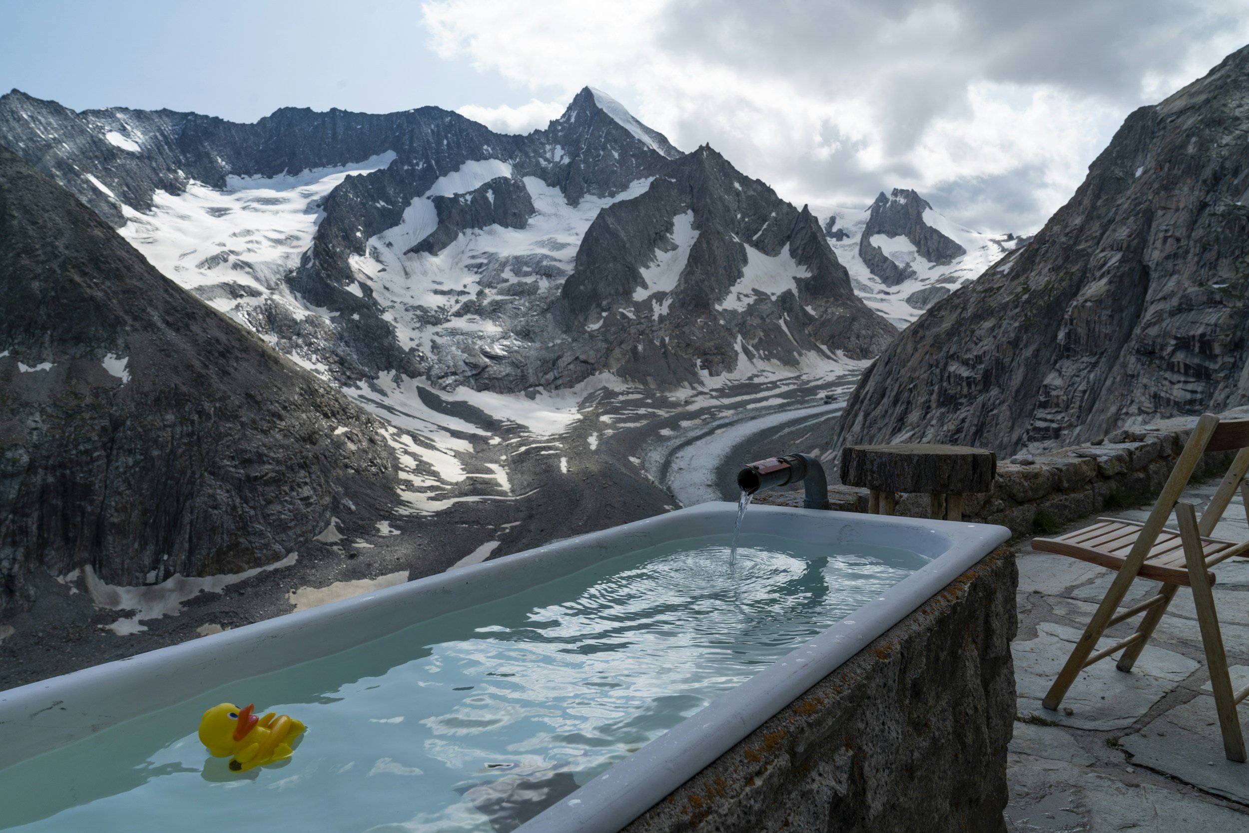 An outdoor hot tub with water, a yellow rubber duck, a mountain range with snow and glaciers in the background, and cloudy sky overhead.