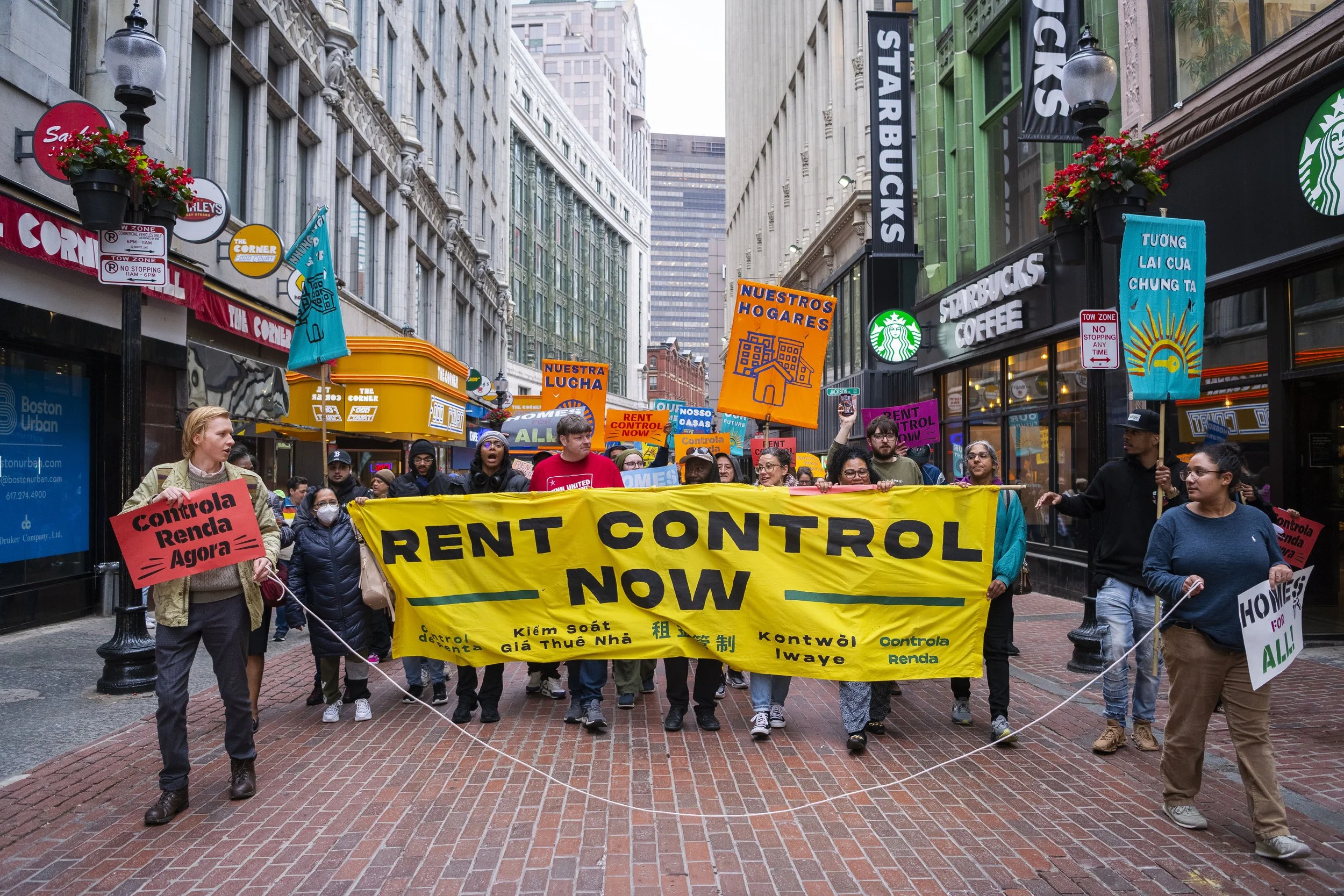 People participating in a protest march on a city street, holding signs and banners advocating for rent control and affordable housing. The march includes diverse individuals walking beneath buildings with storefronts such as Starbucks and various signs in different languages.