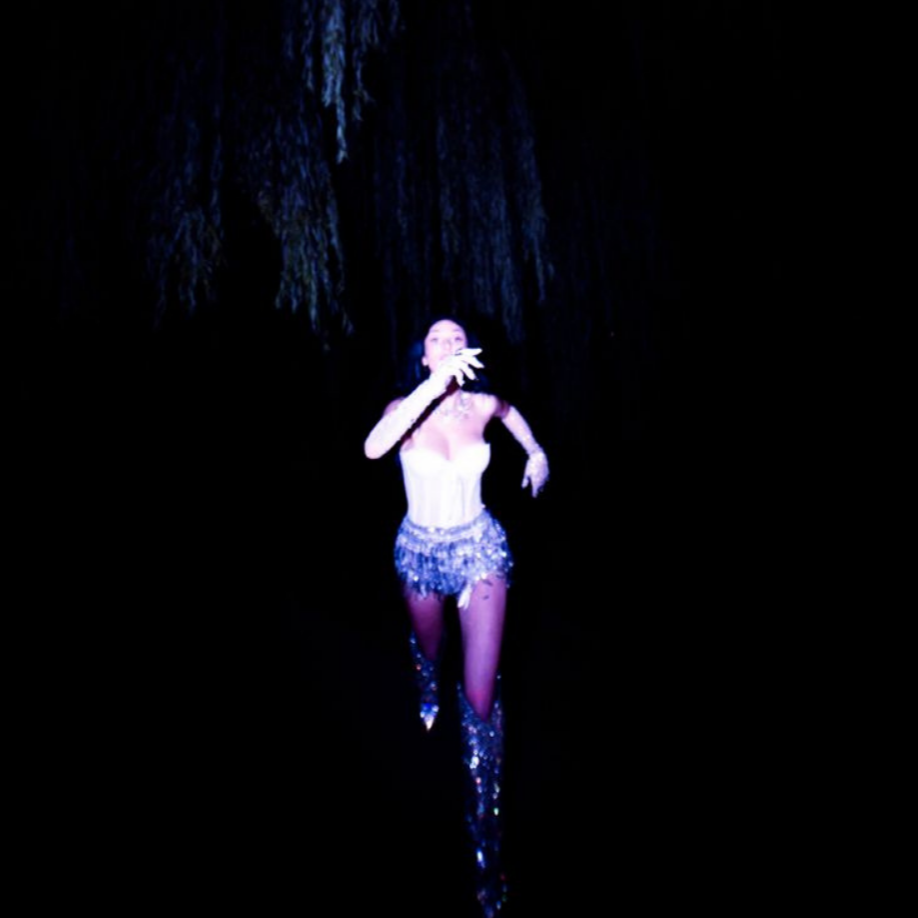 Performer on stage in a sparkling costume with purple tights and high boots, singing or speaking into a microphone, illuminated by spotlight with a dark background.