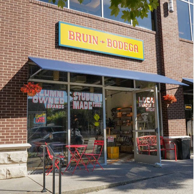 Front exterior of a store named 'Bruin Bodega' with a blue awning, red patio furniture, and hanging flower baskets, with a glass door showing the interior.