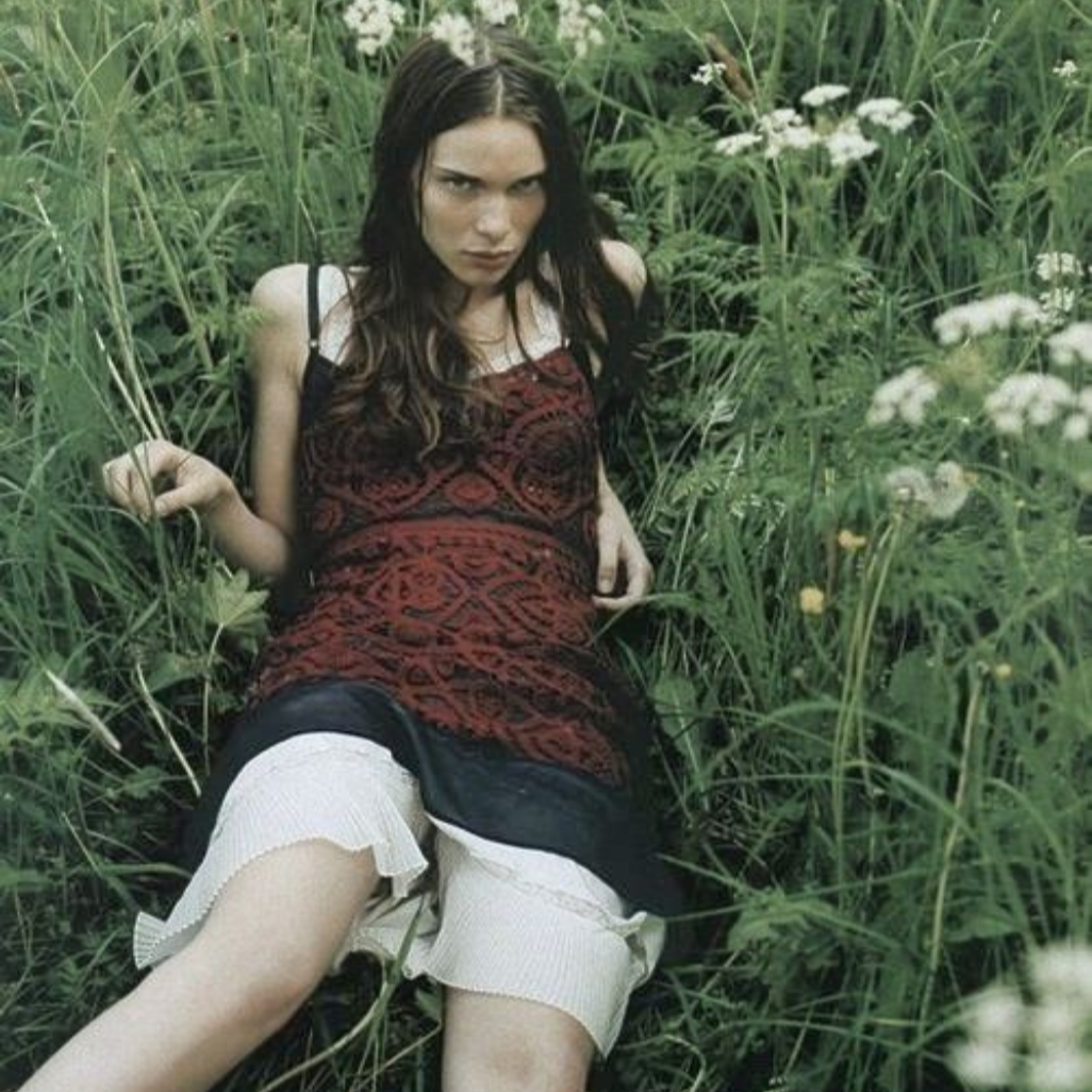 A young woman with long dark hair and wearing a patterned dress is sitting in tall green grass amidst wildflowers, looking directly at the camera.