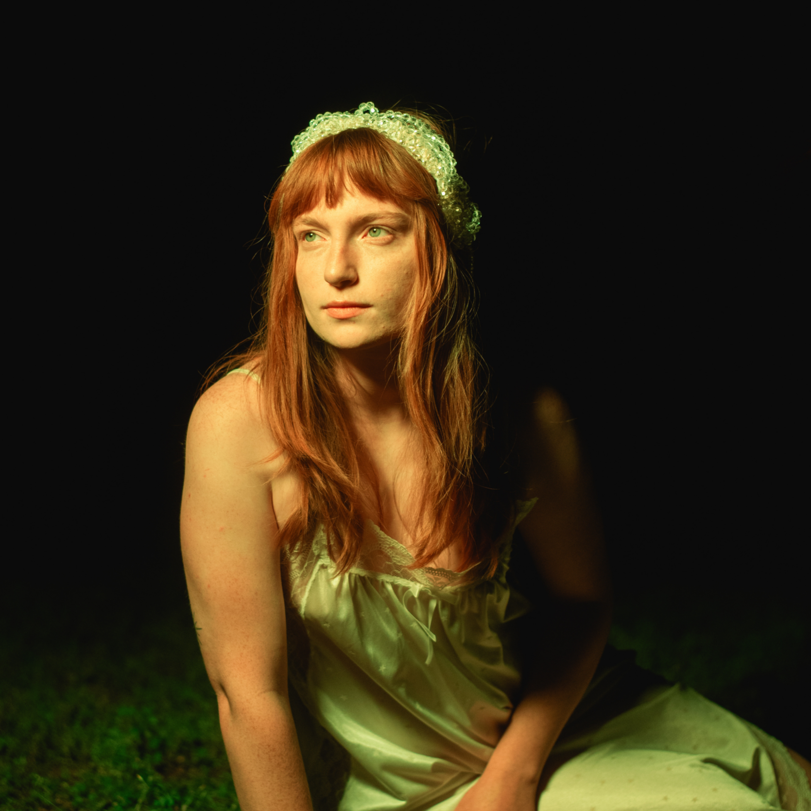 A young woman with long red hair and green eyes sitting outdoors at night, wearing a cream-colored satin dress with lace details and a beaded headband, illuminated by soft light.