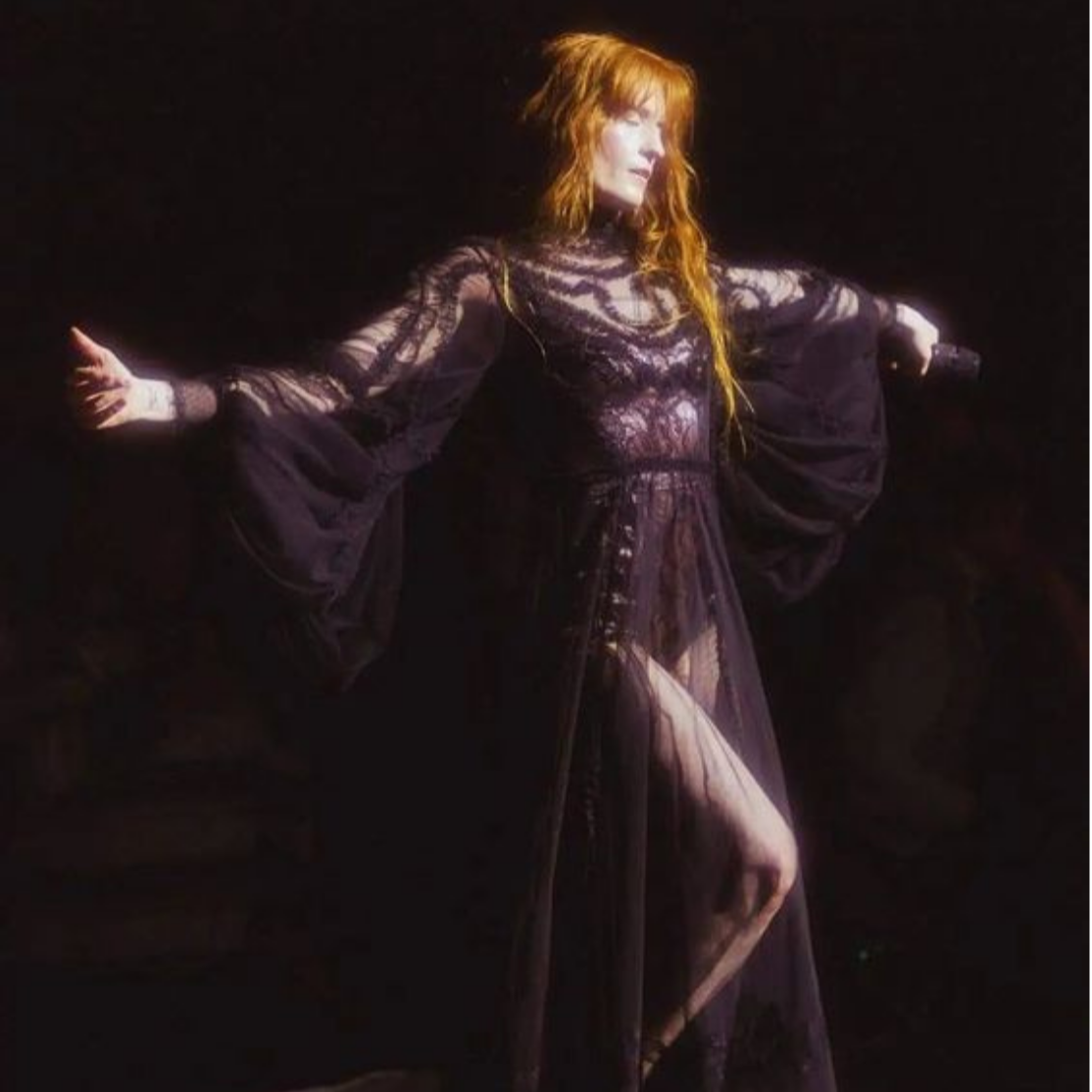 A woman with long red hair wearing a black, sheer, lace dress with puffy sleeves, standing with arms extended against a dark background.