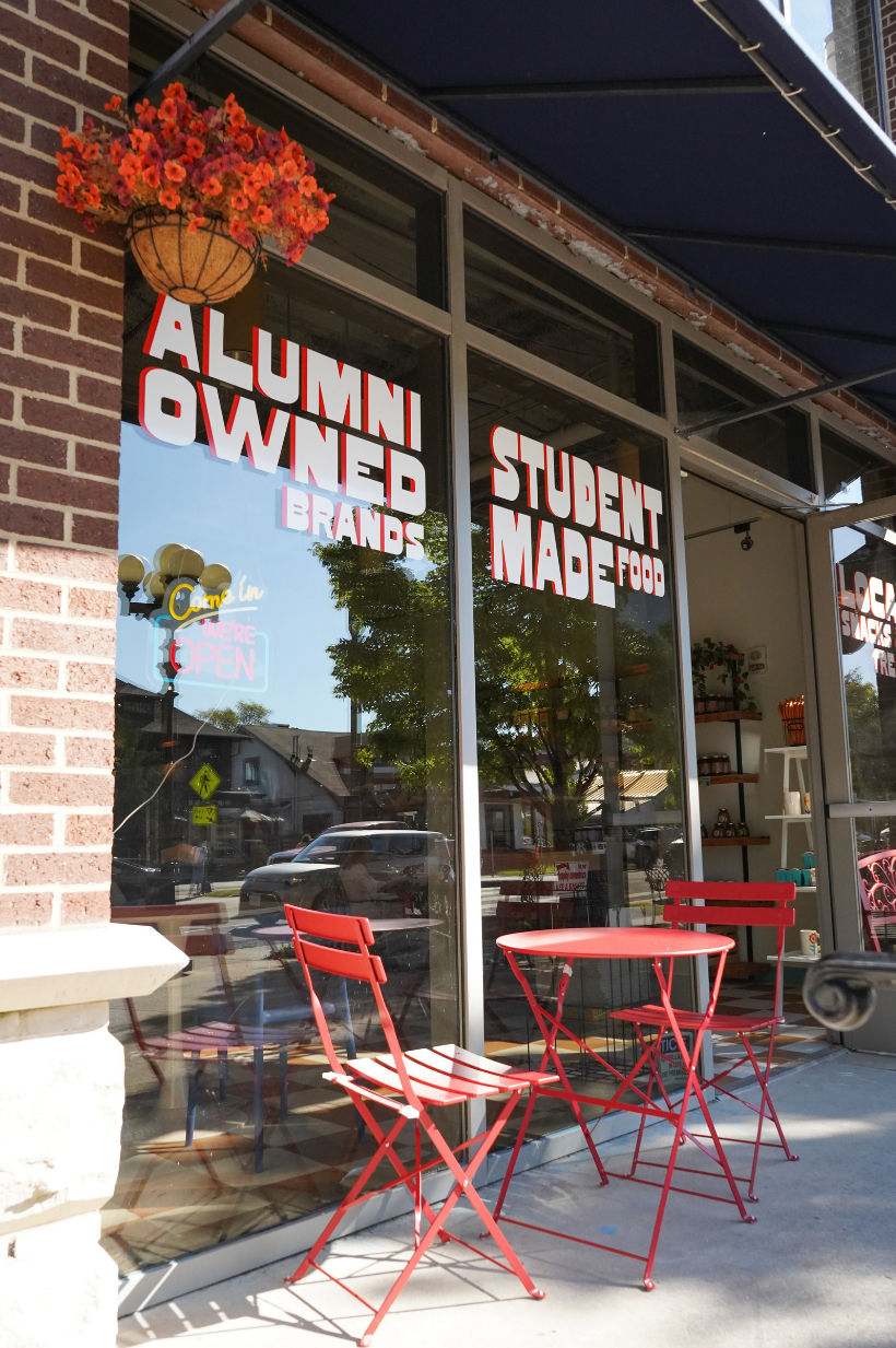 A storefront with large window displaying signs that read 'Alumni Owned Brands' and 'Student Made Food.' Outside, there are red metal chairs and a table on the sidewalk. A hanging flower basket is above the window, and trees and cars are reflected in the glass.
