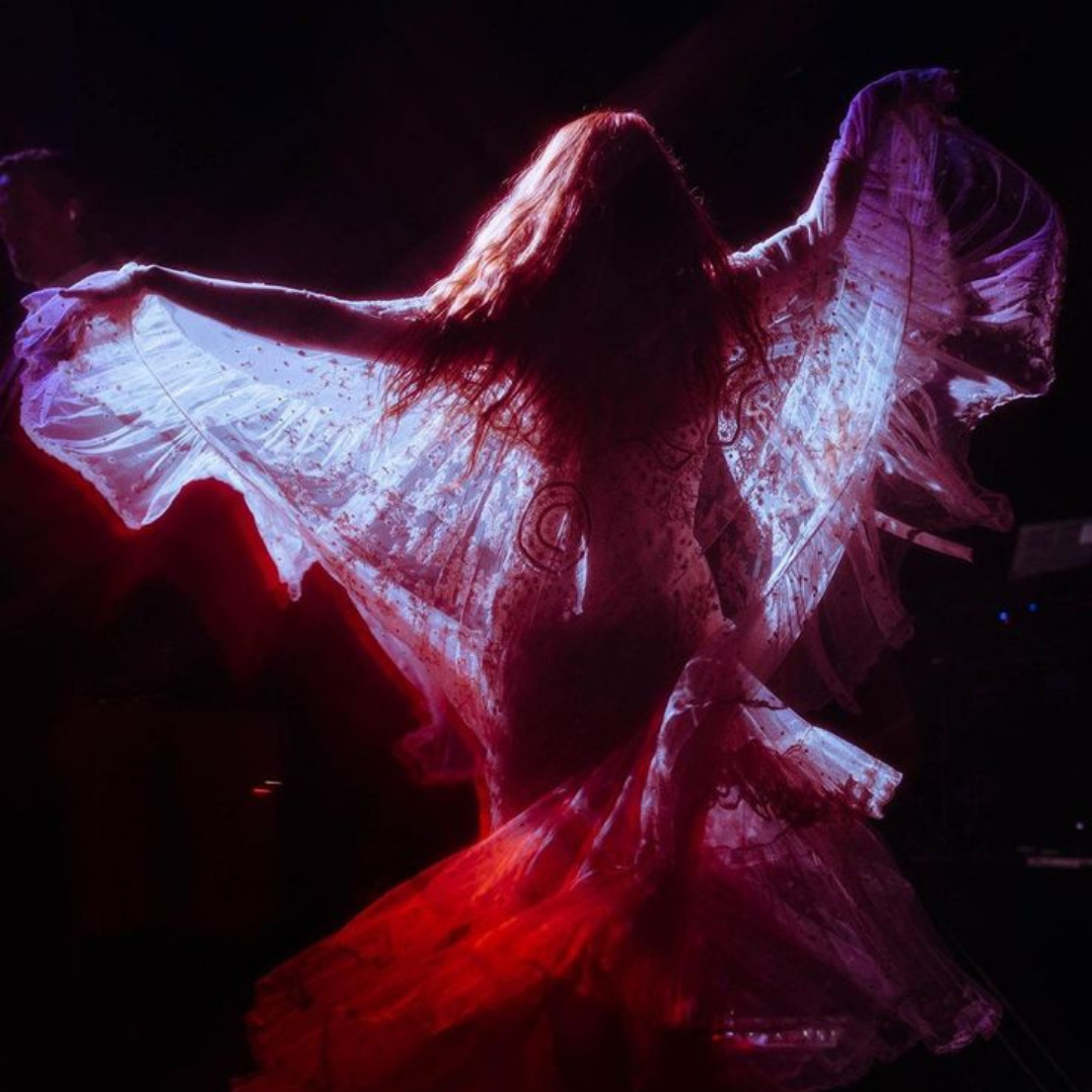 A person with long hair dancing in a dark setting, wearing a flowing, illuminated dress with vibrant pink and purple lights.