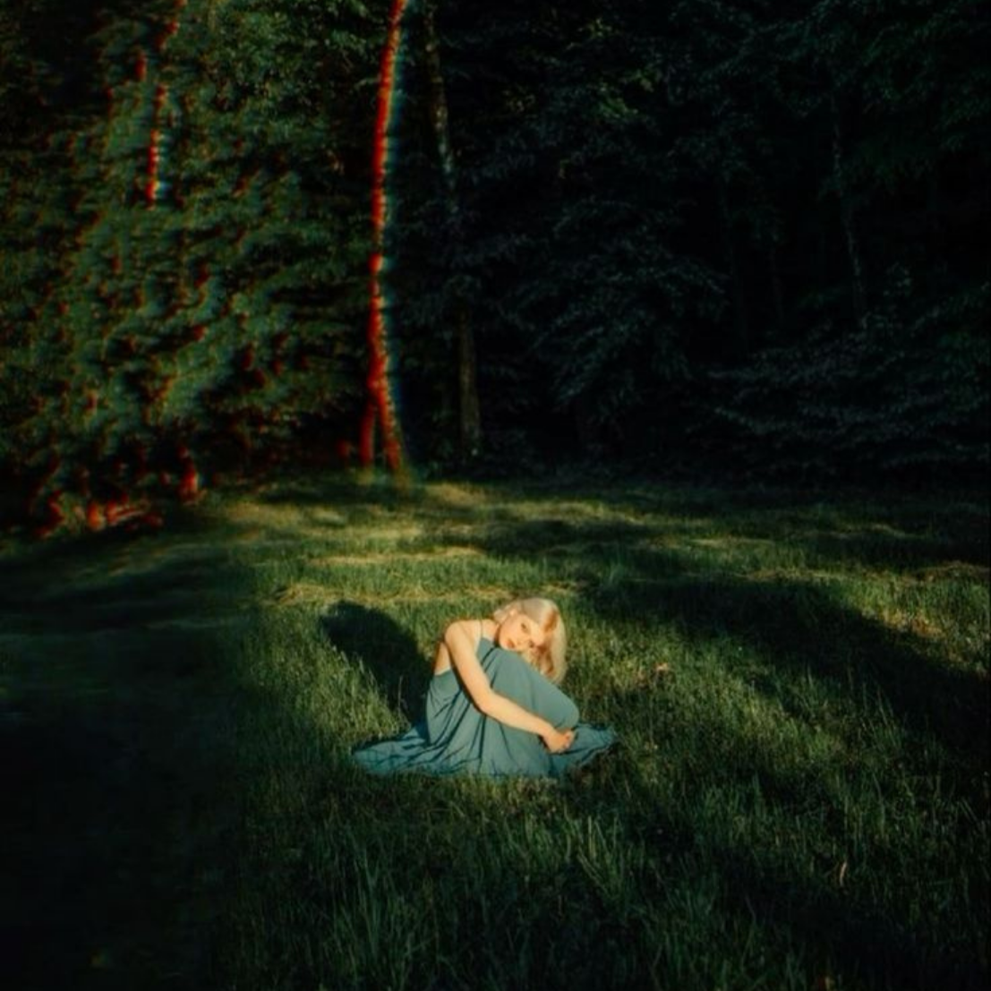 A woman with blonde hair sitting on the grass in a forest clearing, wearing a blue dress and hugging her knees, with trees and shadows around her.