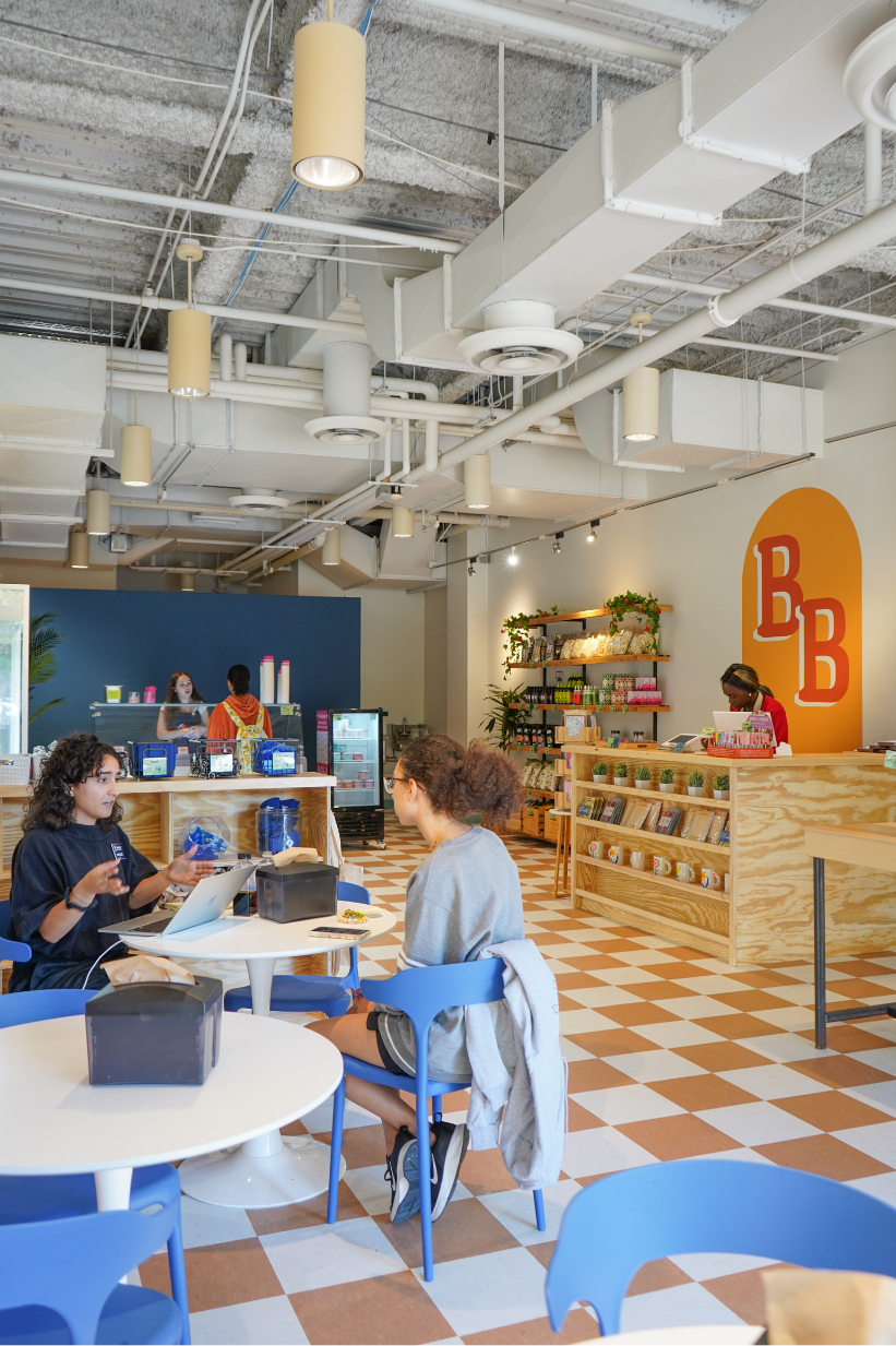 A café with two women sitting at a white round table, talking. The café has a checkered floor, exposed ceiling pipes, and a counter at the back with a staff member. Shelves display snacks and drinks, and there's a colorful orange logo on the wall.
