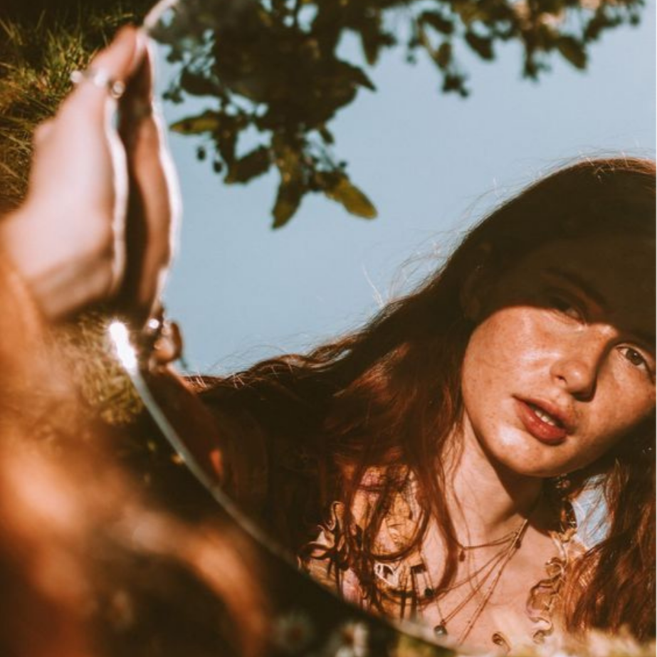 A woman with red hair looking into a reflective surface, possibly a mirror, with water and trees in the background.