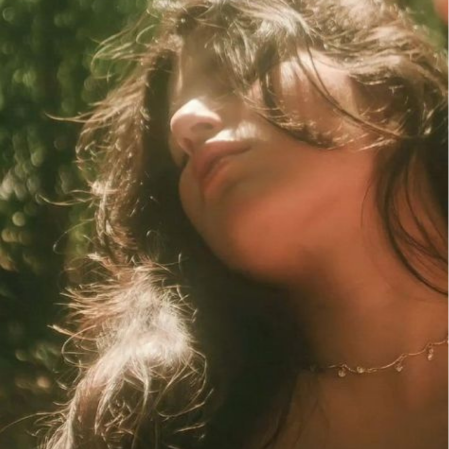 Close-up of a woman's face, with her hair partially covering her face, looking upward outdoors with greenery in the background.