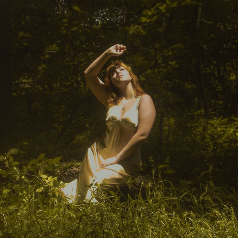 A woman with curly red hair sitting on a log in a forest, wearing a satin dress, looking up with her hand raised above her head.