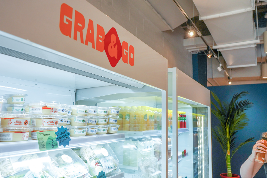 Section of a grocery store called 'Grab & Go' with refrigerated items including hummus, bottled drinks, and a person's hand holding a beverage on the right, and a potted plant in the background.