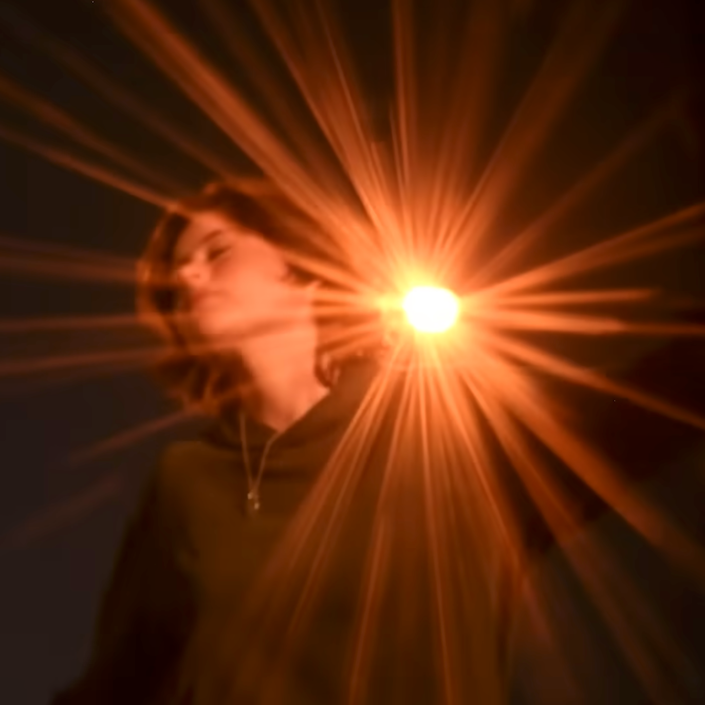 Person with closed eyes and a slight smile, face illuminated by a bright, radiant light emitting rays in all directions, set against a dark background.