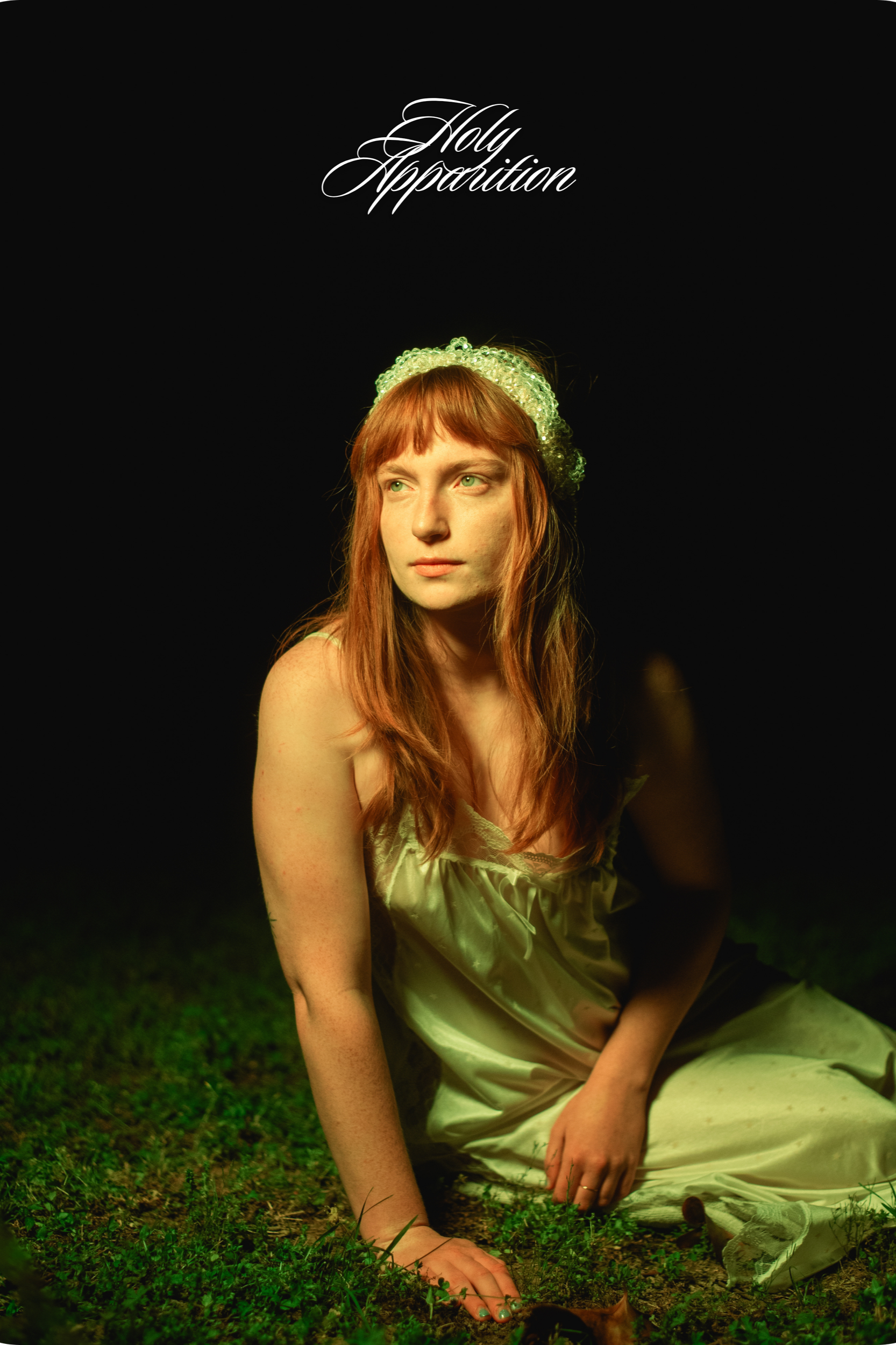 A young woman with red hair, green eyes, wearing a beige satin dress and a beaded headband, is sitting outdoors on green grass at night. The background is dark, and she is looking to her right, reaching her hand towards a small animal or object on the ground.