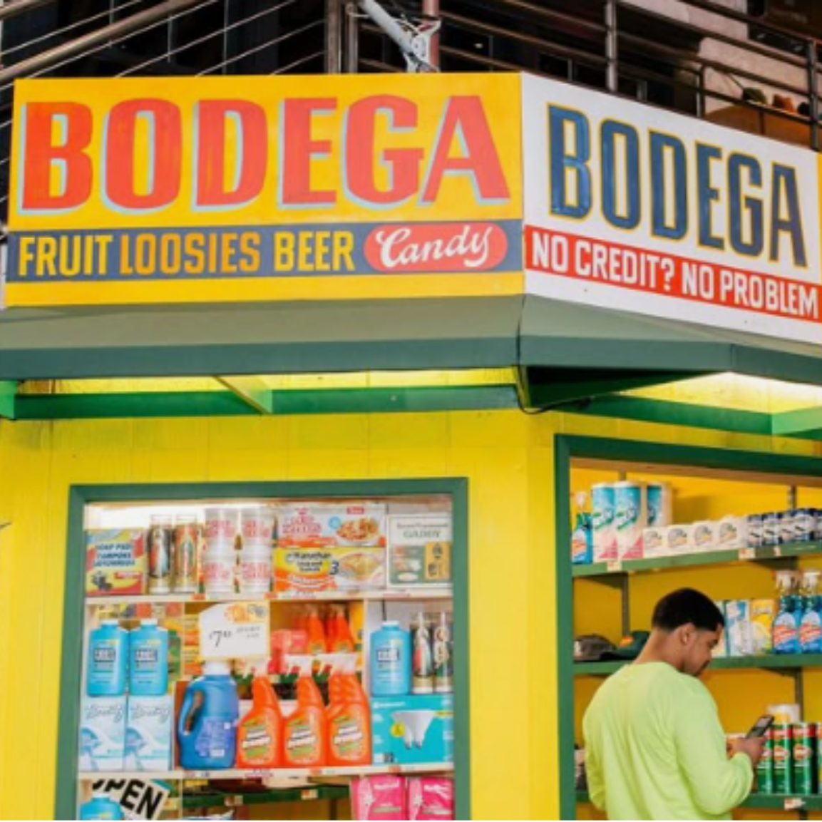 A bright yellow storefront with a sign reading 'BODEGA' and advertisements for fruit looses, beer, and candy. A man in a neon green shirt is shopping inside, looking at products on the shelves.