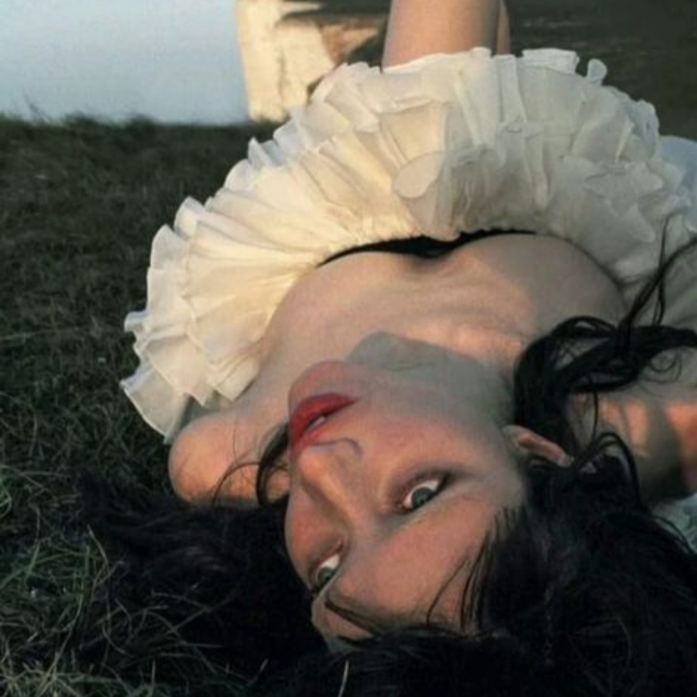 A woman lying on grass, wearing a white frilled dress, with long dark hair, looking up at the camera.