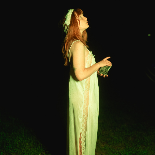 A woman with red hair, wearing a light green dress and a hat, standing outdoors at night and holding a small box.