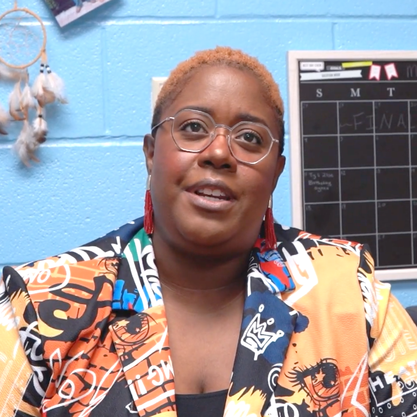 A woman with short, dyed orange hair and glasses, wearing a colorful, patterned jacket, smiling in front of a blue wall with a dreamcatcher and a calendar board.