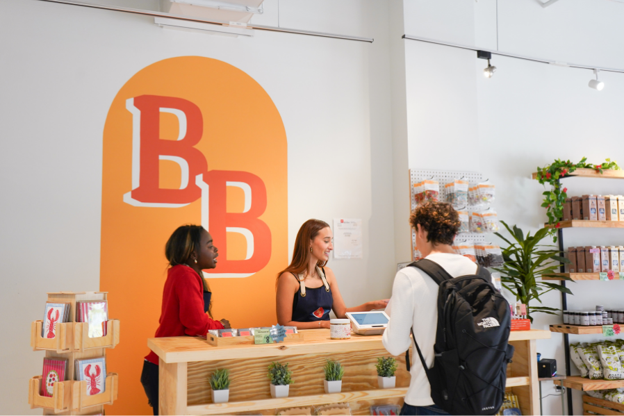 A store clerk and a customer at the checkout counter in a retail store with a large orange "B" logo on the wall behind them.