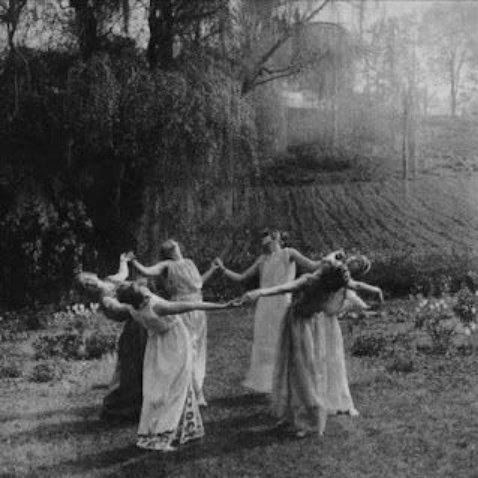Four women dancing outdoors holding hands in a circle, wearing long dresses, in a rural setting with trees and fields in the background.