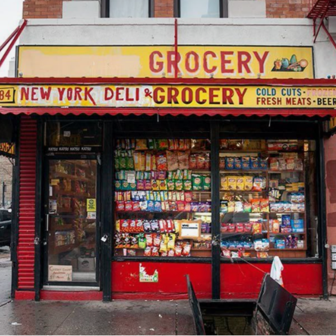 storefront with a yellow and red sign reading 'GROCERY' and 'NEW YORK DELI & GROCERY,' displaying snacks and packaged foods inside.