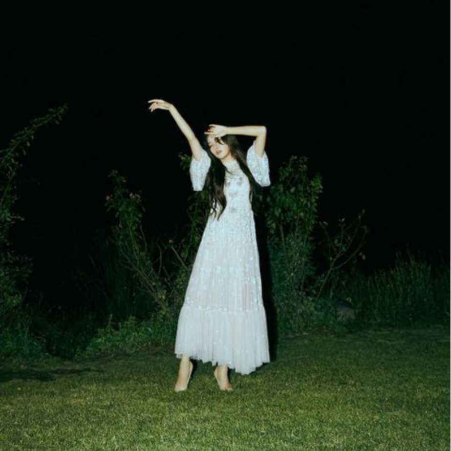 A woman in a long white dress standing on grass at night, with her arms raised above her head and a dark background with some greenery.
