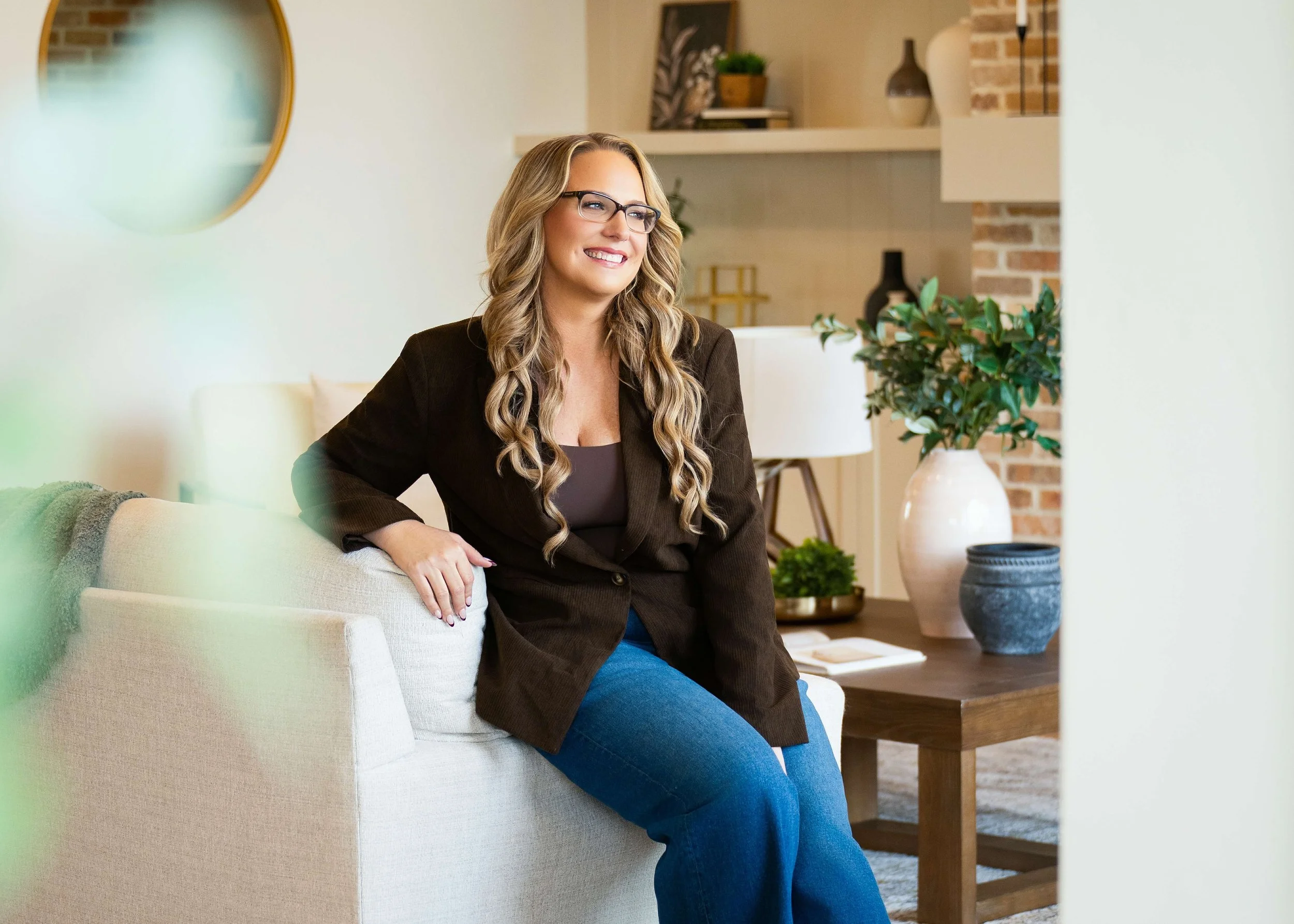A woman with long curled hair, glasses, a black blazer, and blue jeans sitting on a beige sofa in a modern living room. She is smiling and looking off to the side, with plants and decorative items in the background.