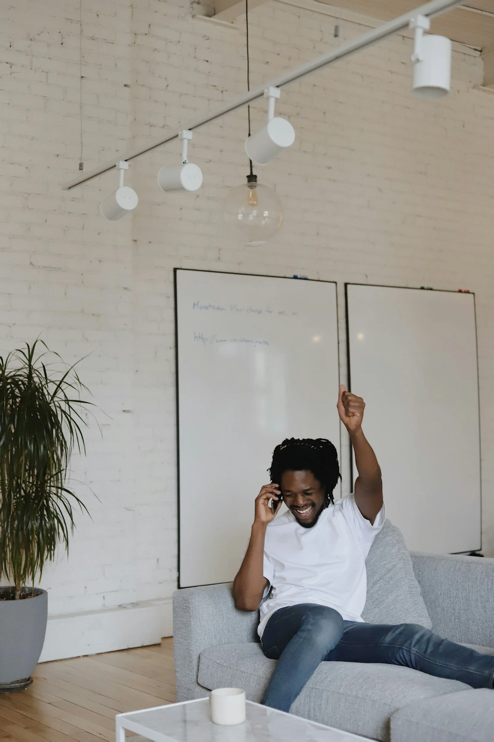 A man sitting on a gray sofa in a modern, minimalist room, talking on a phone with a big smile, raising his fist in celebration or excitement. The room has a white brick wall, a whiteboard, a potted plant, and a contemporary light fixture hanging from the ceiling.