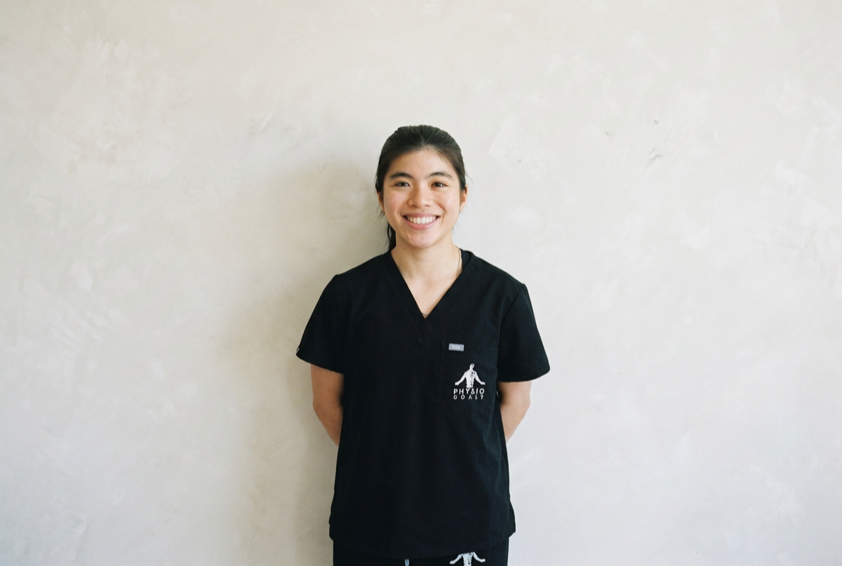 A young woman in black medical scrubs standing against a plain light-colored wall, smiling with hands behind her back.