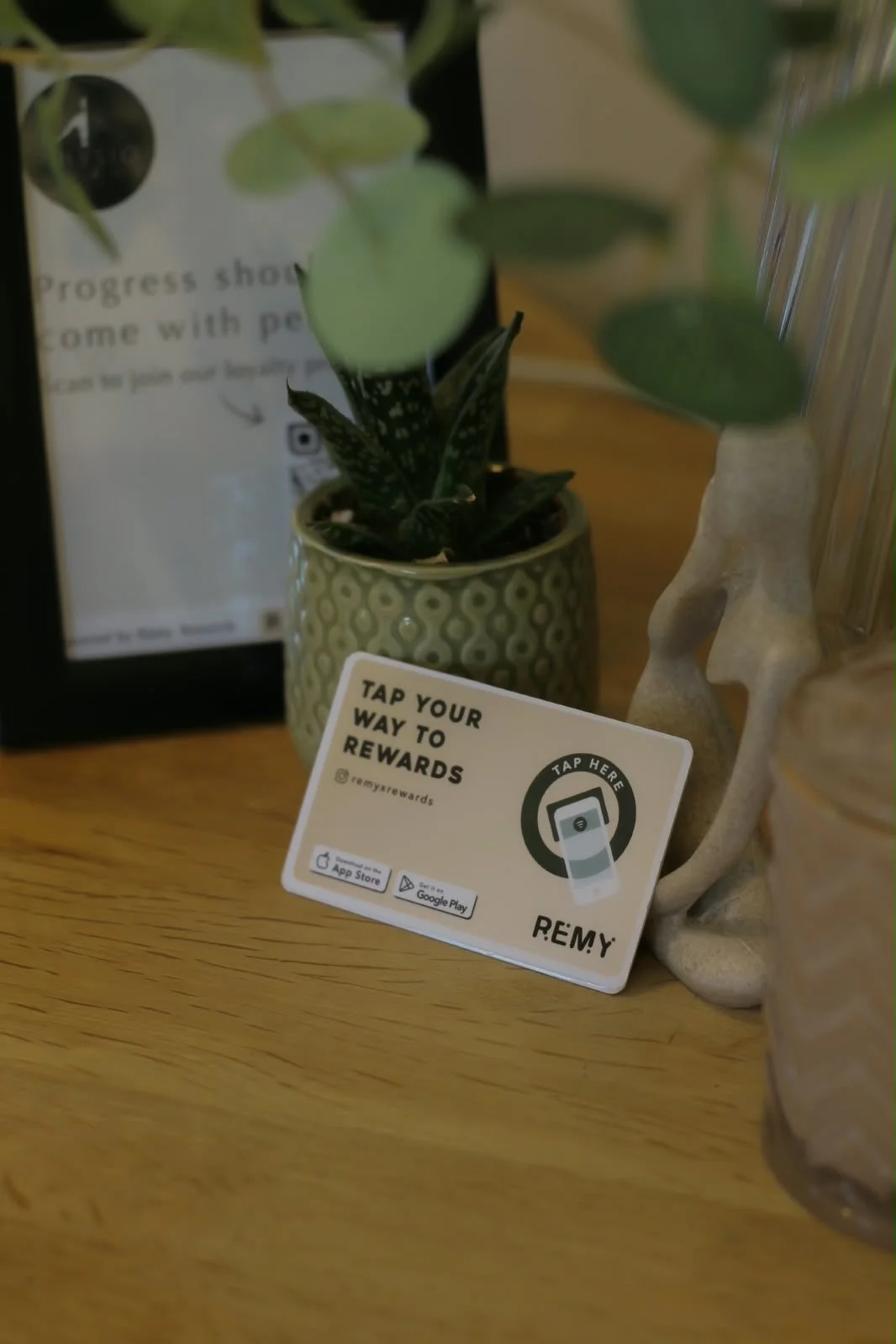 A small plant in a decorative green pot, a promotional card for REMY rewards, promoting Physio Goals loyalty programme, with an illustration of a phone and a 'Tap Here' icon, and part of a human face sculpture, on a wooden surface.