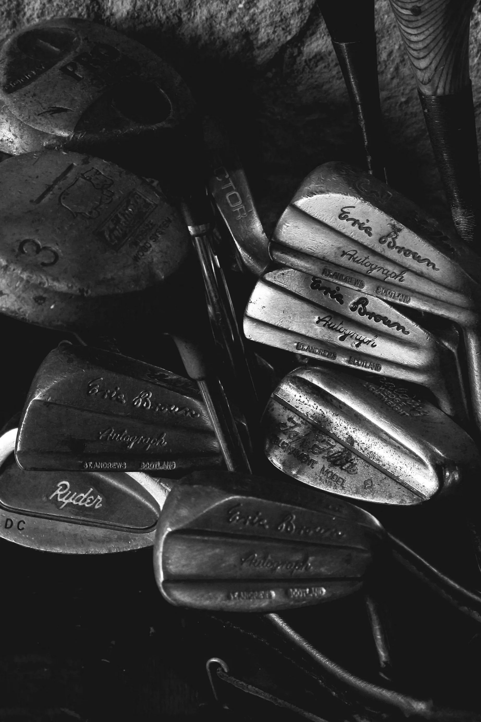 A collection of vintage golf clubs with various brands and models, including Eric Brown, Ryder, and PING, placed together in a seemingly random arrangement.
