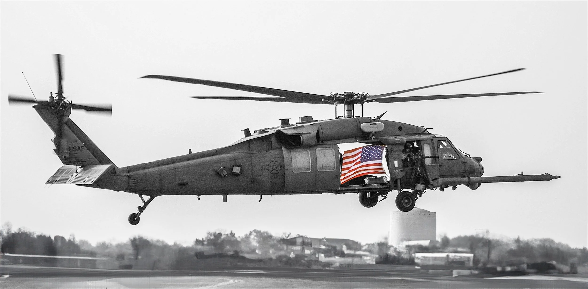 A military helicopter in mid-air with an American flag decal on its side, flying over a landscape with trees and structures in the background.