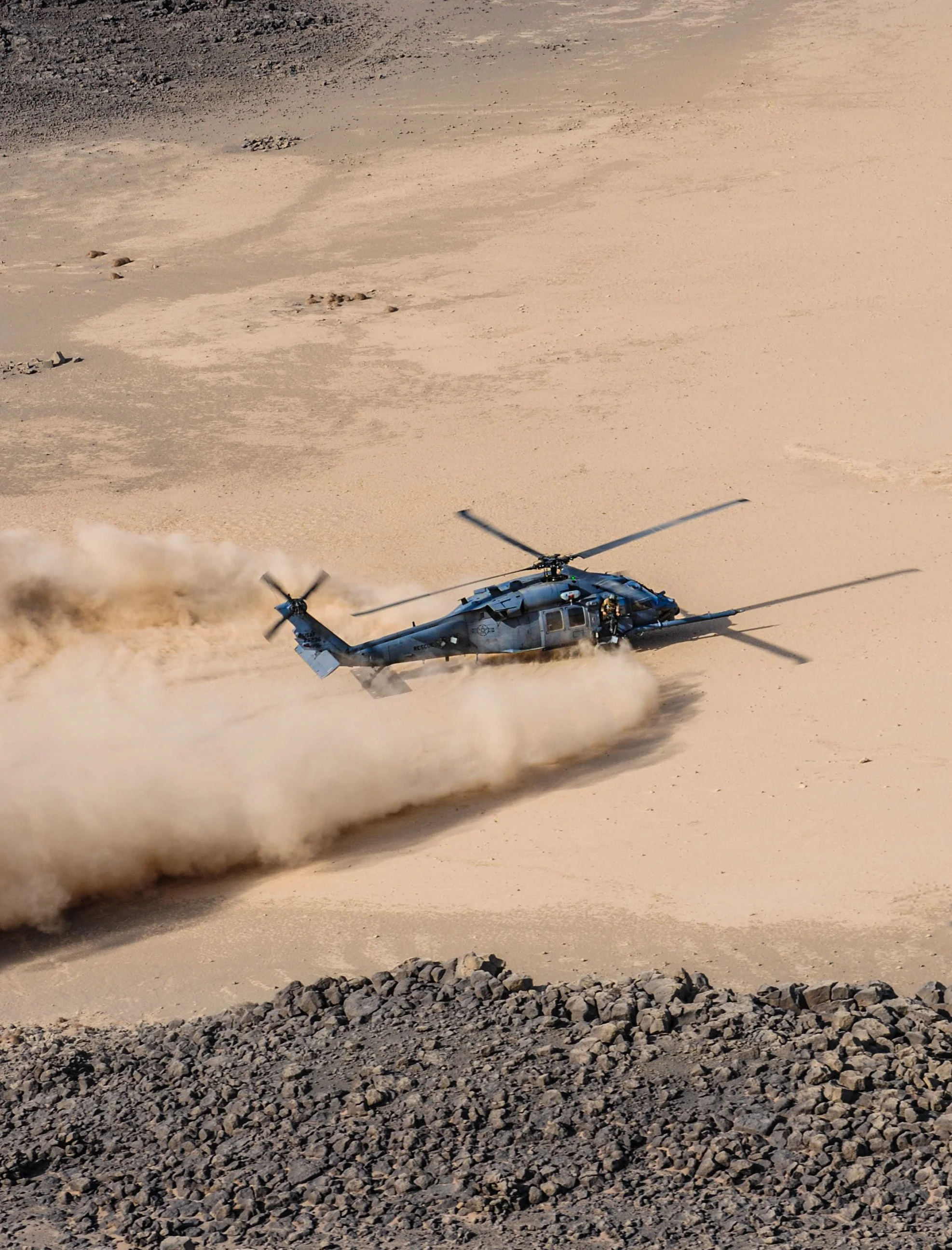 A military helicopter flying low over a desert landscape, kicking up dust and sand.
