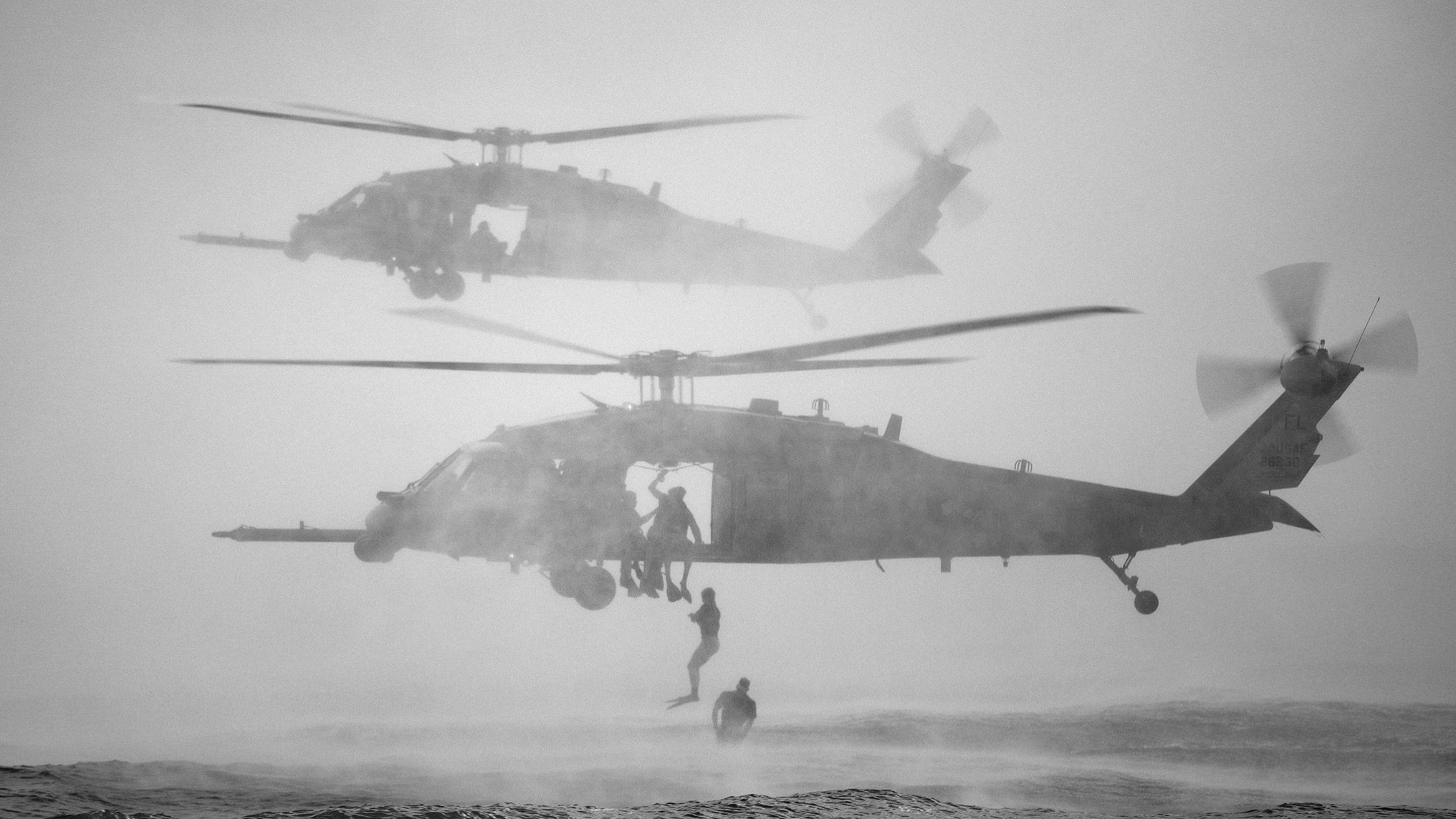 Two military helicopters flying close to the ground with military personnel hanging underneath, some on the ground, in a hazy environment.