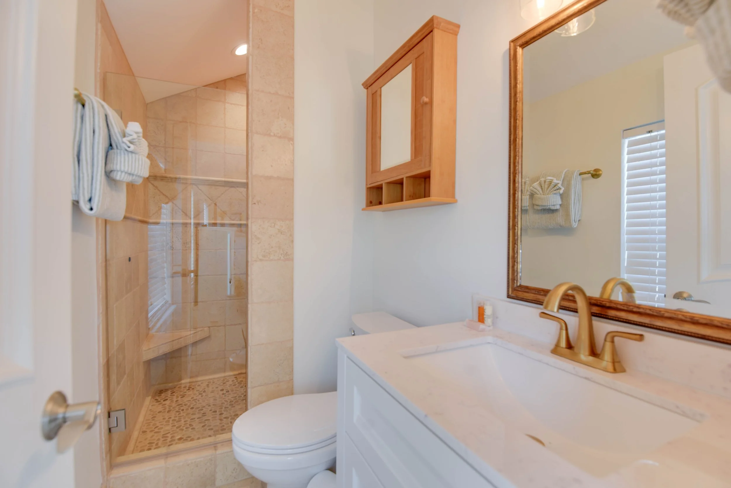 Bathroom with a wooden cabinet, a marble countertop, a large mirror, a white toilet, and a glass-enclosed shower with beige tiles.
