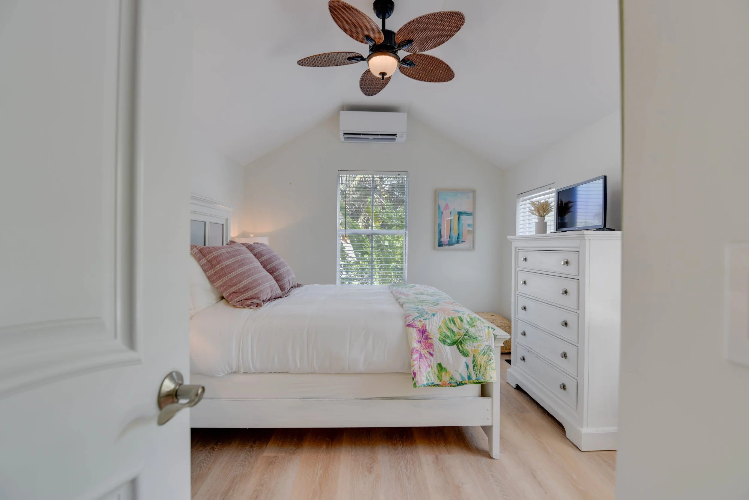 View of a bedroom through an open door, featuring a bed with pink striped pillows, a colorful bedspread, a white dresser with a TV, and windows showing greenery outside. A ceiling fan with leaf-shaped blades is on the ceiling.