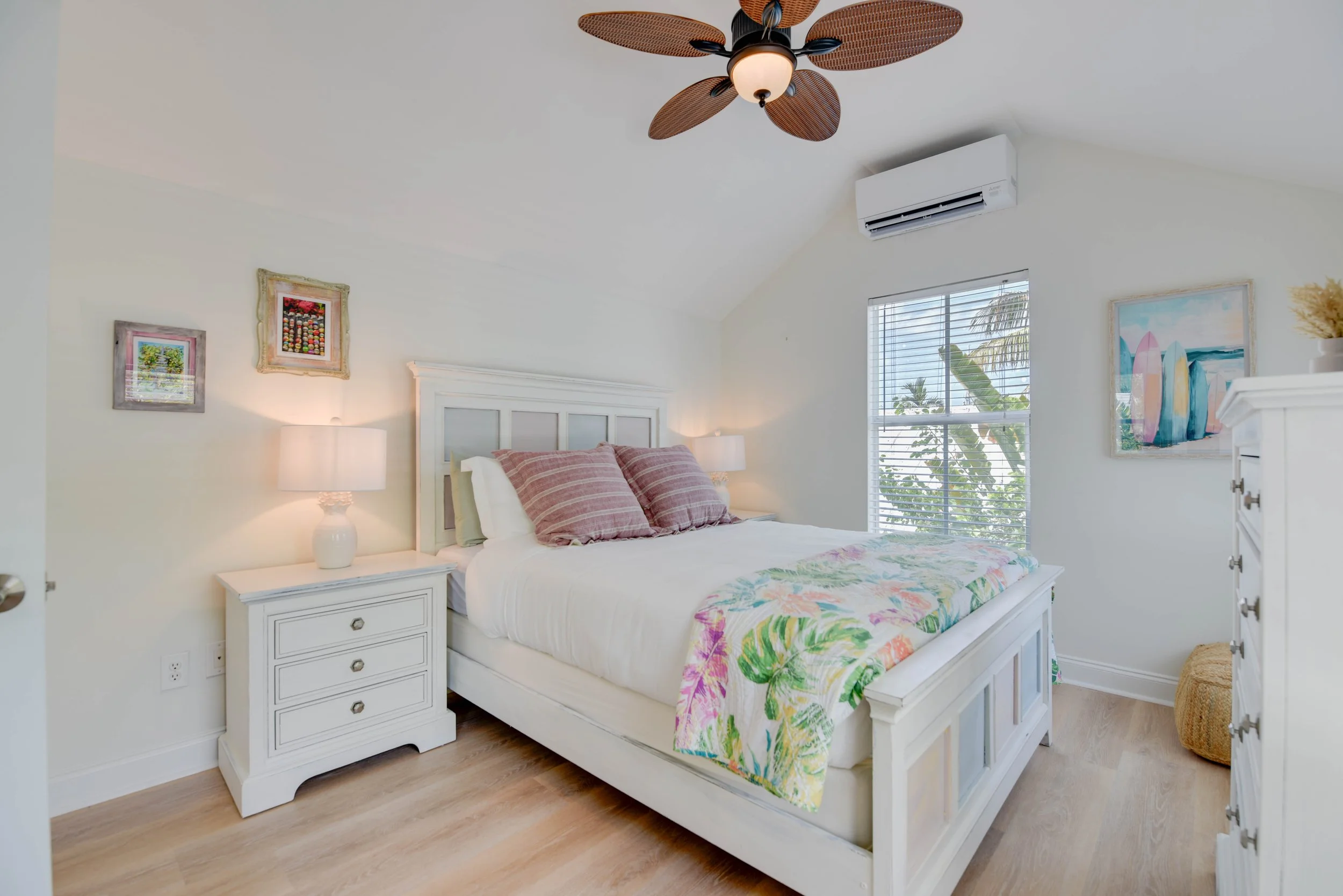 A bedroom with a white wooden bed frame, pink striped pillows, and a colorful floral bedspread. There are two white nightstands with lamps, colorful framed artwork on the walls, a window with blinds showing tropical trees outside, a ceiling fan with 
