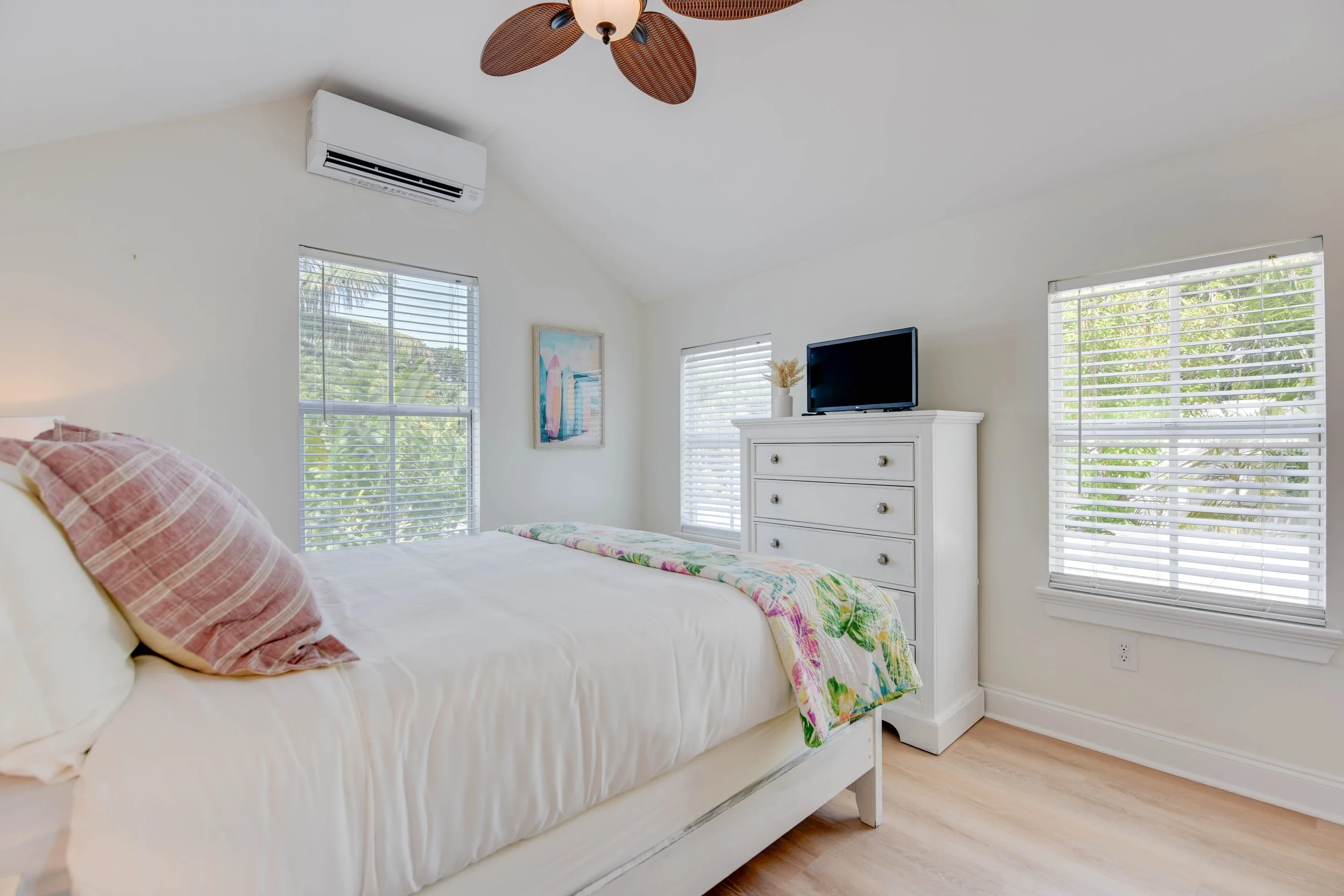 A bedroom with white walls, a bed with pink and white striped pillows, a colorful blanket, windows with white blinds, a white dresser with a small TV and a vase, framed artwork, and a ceiling fan with leaf-shaped blades.