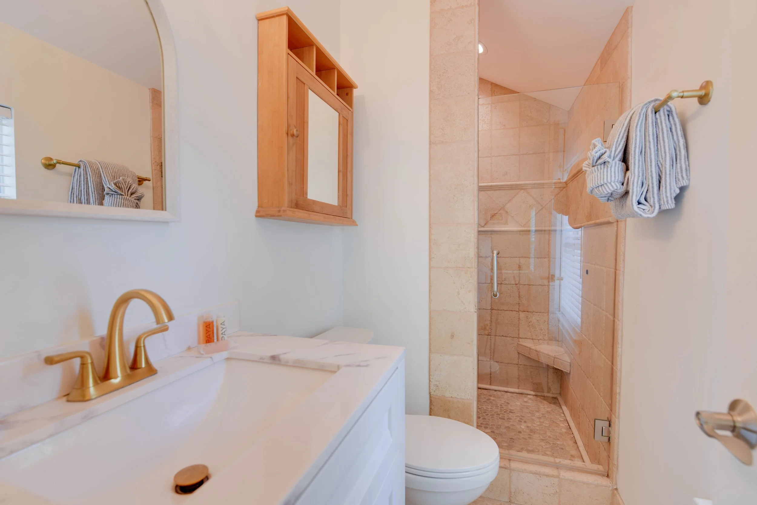 Bathroom with a white vanity and gold fixtures, beige tiled shower area with a glass door, towels hanging on a gold towel rod, and a mirror above the sink.