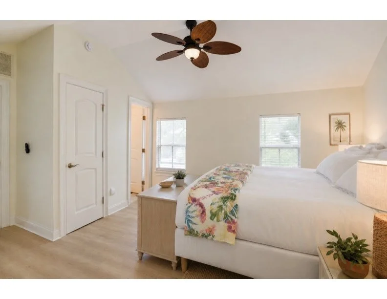 Bedroom with white walls, wooden floor, a large bed with white bedding, a colorful floral blanket, two windows, a nightstand with a plant, and a ceiling fan with wooden blades.