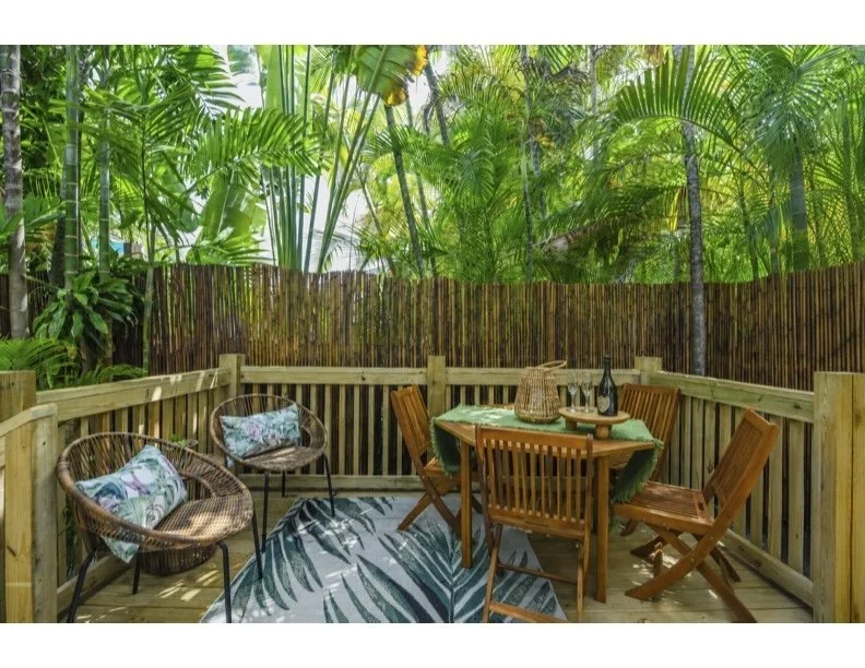 Outdoor wooden deck with seating area, surrounded by tall tropical plants and bamboo fence