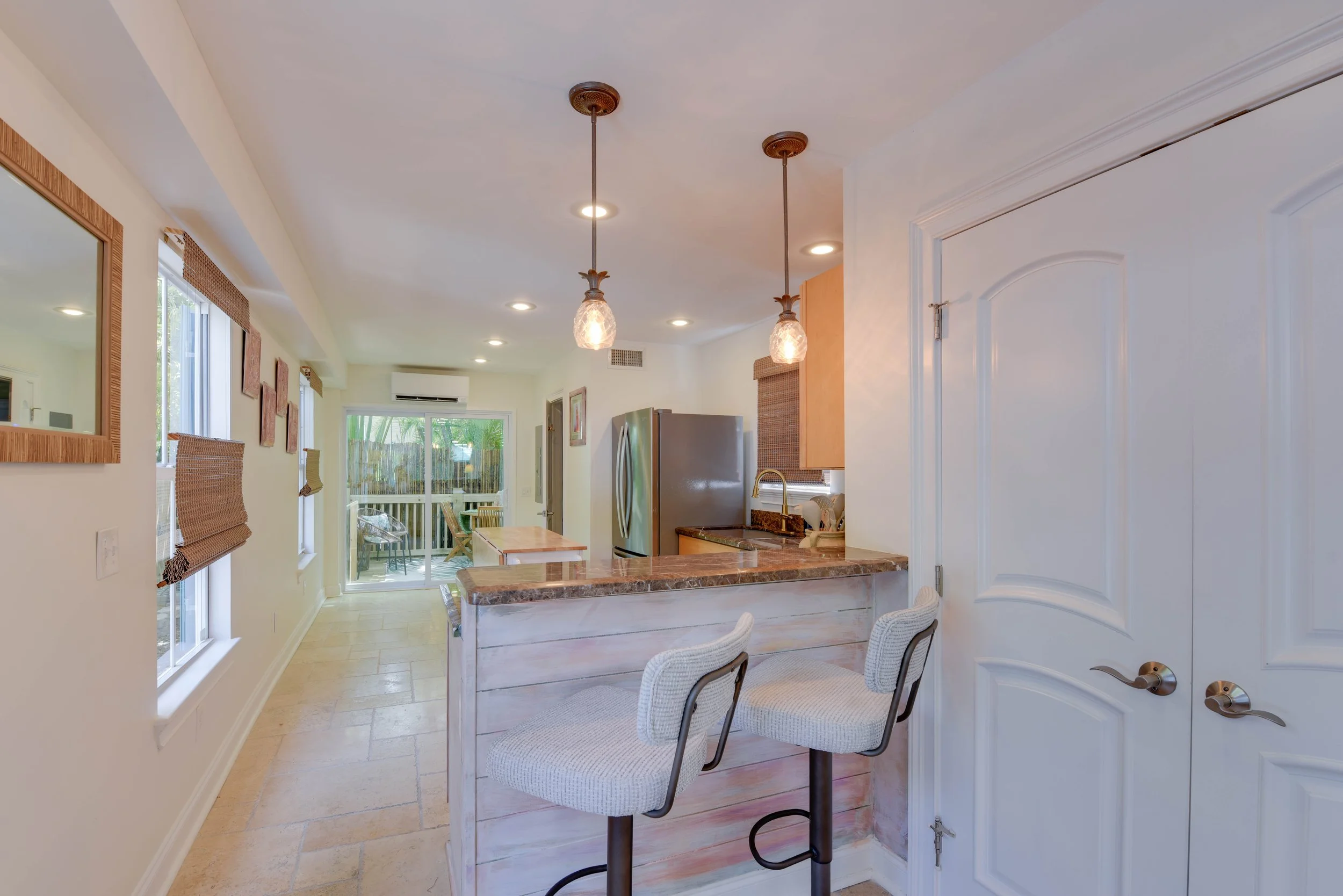 A bright kitchen with pendant lights above a marble counter, with windows and a sliding door in the background, and dining chairs around a small table.