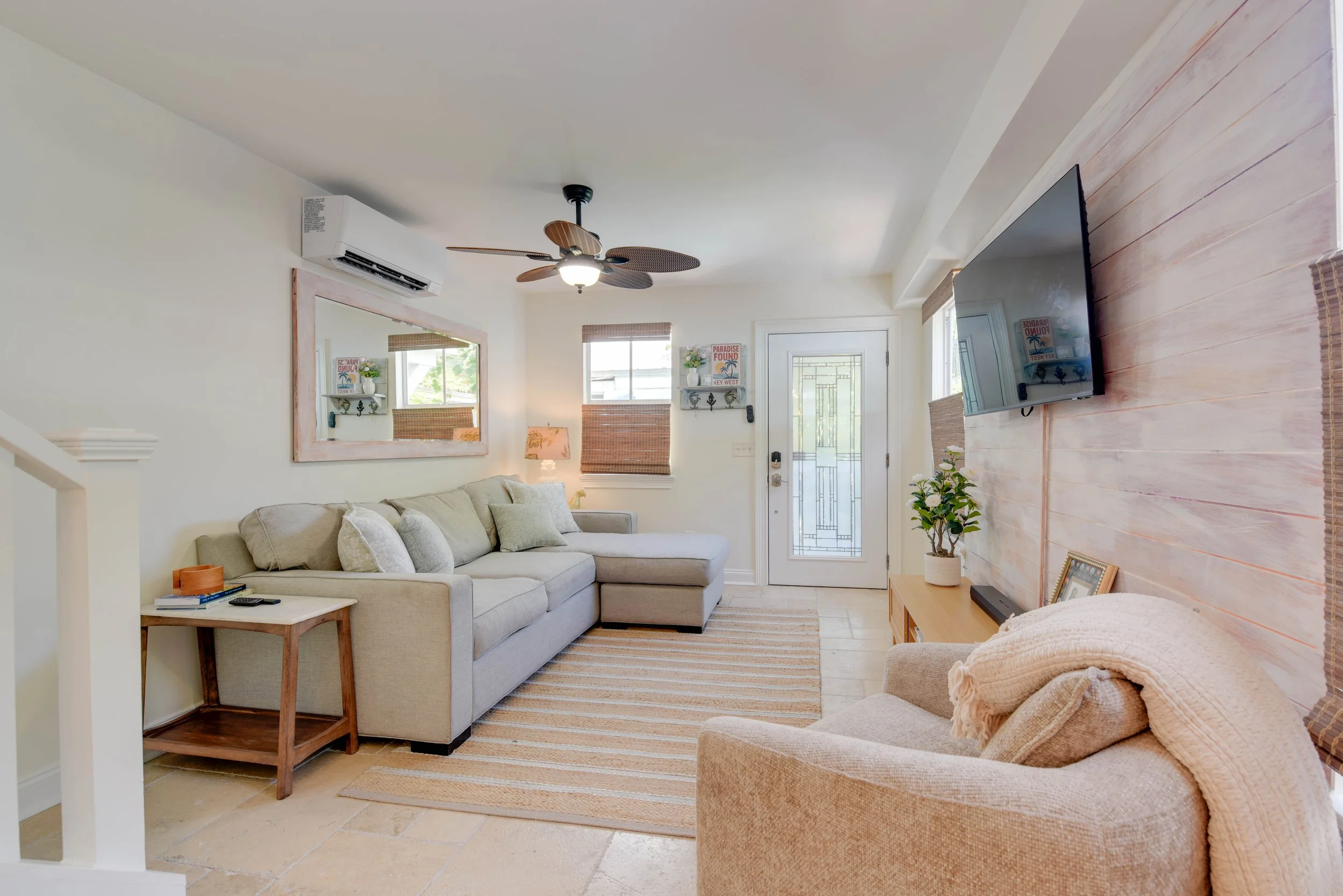 Living room with beige couch, side table, wall-mounted TV, ceiling fan, windows with wicker blinds, and decorative wall shelves