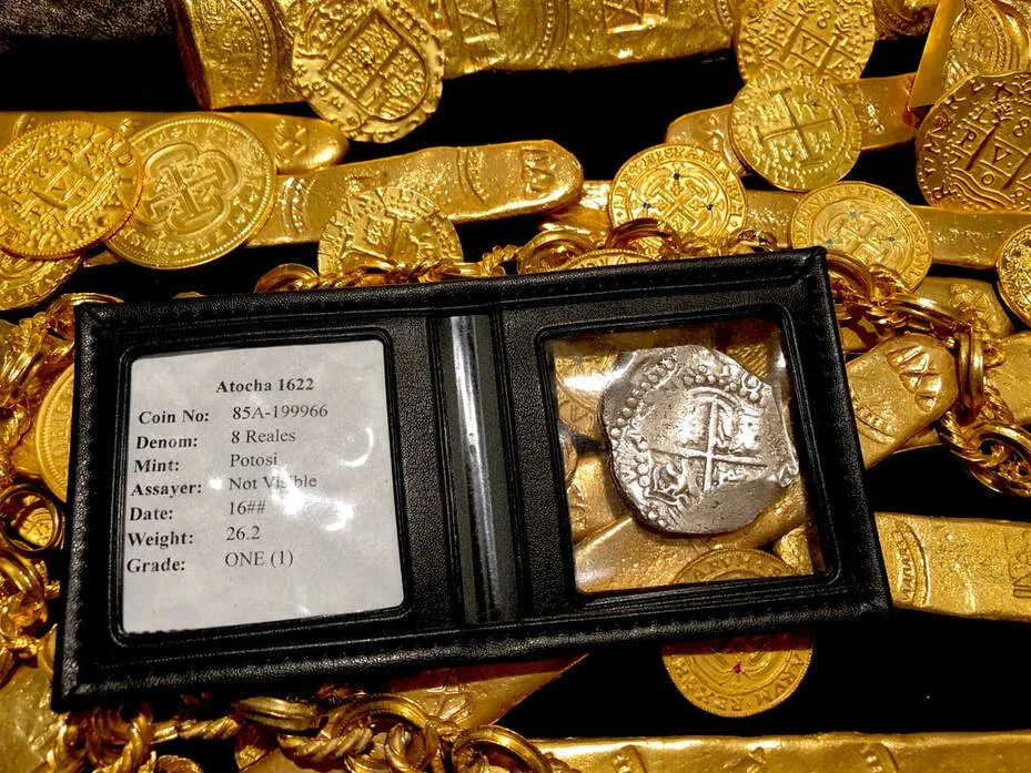 A display of gold coins and jewelry with a small black case containing a silver coin labeled 'Atocha 1622, 8 Reales, Potosi, not visible assayer, weight 26.2, grade ONE.'