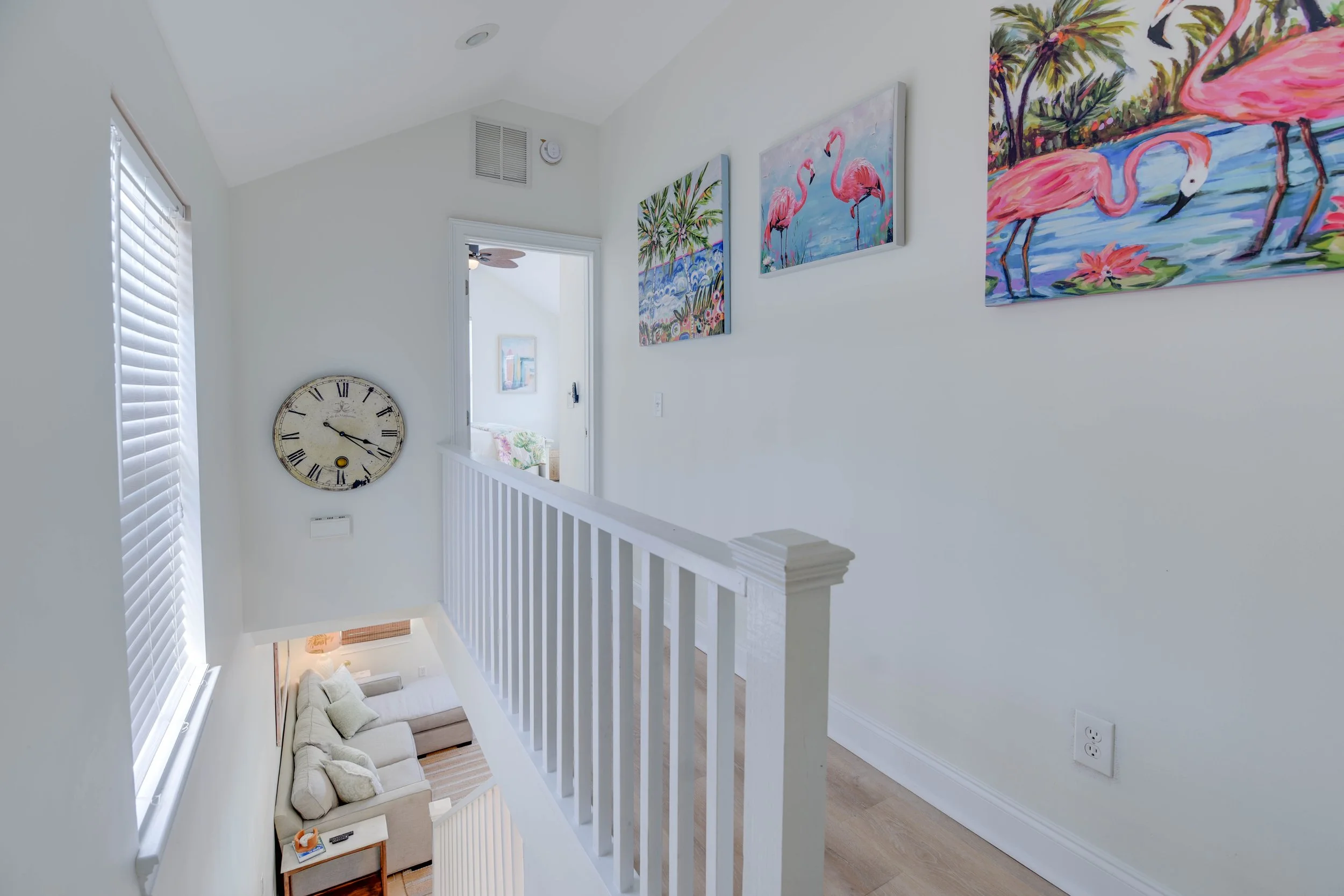 View of a second-floor hallway with white walls, three colorful paintings of flamingos and tropical scenery, a large vintage clock, and a view into a living room with a beige sofa.