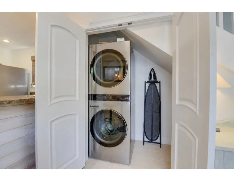 A stacked washer and dryer unit in a laundry closet behind open double doors, with an ironing board to the right.