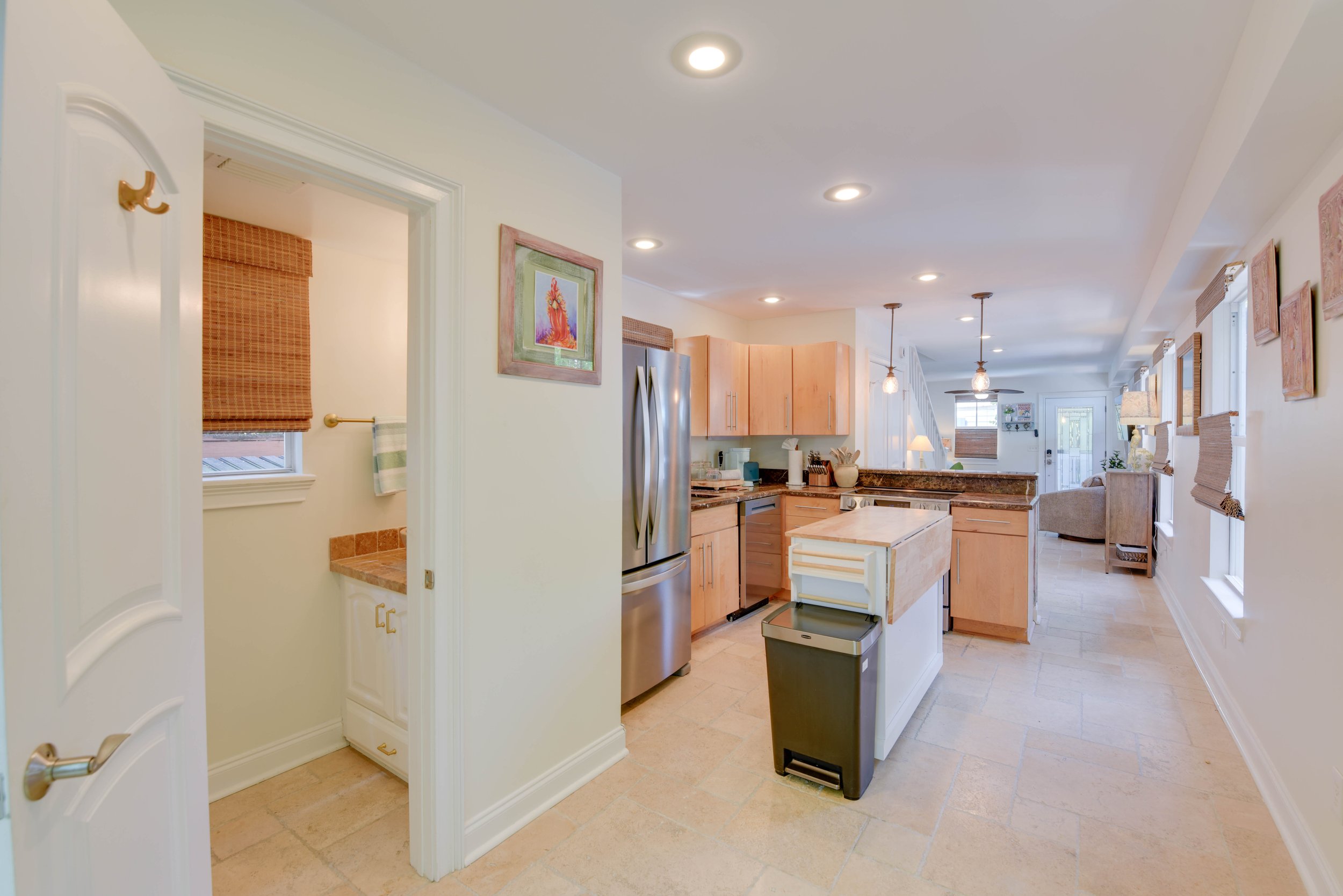 Interior view of a kitchen with wooden cabinets, stainless steel refrigerator, granite countertops, and pendant lighting hanging from the ceiling. There are windows with woven blinds, and artwork on the walls.